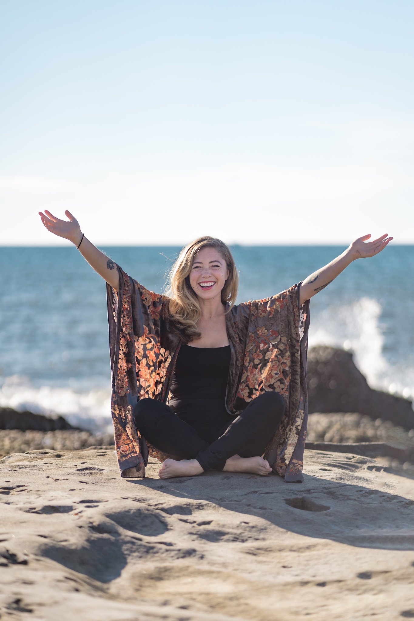 Woman sitting on sand with arms raised, smiling, near the ocean with rocks in the background.
