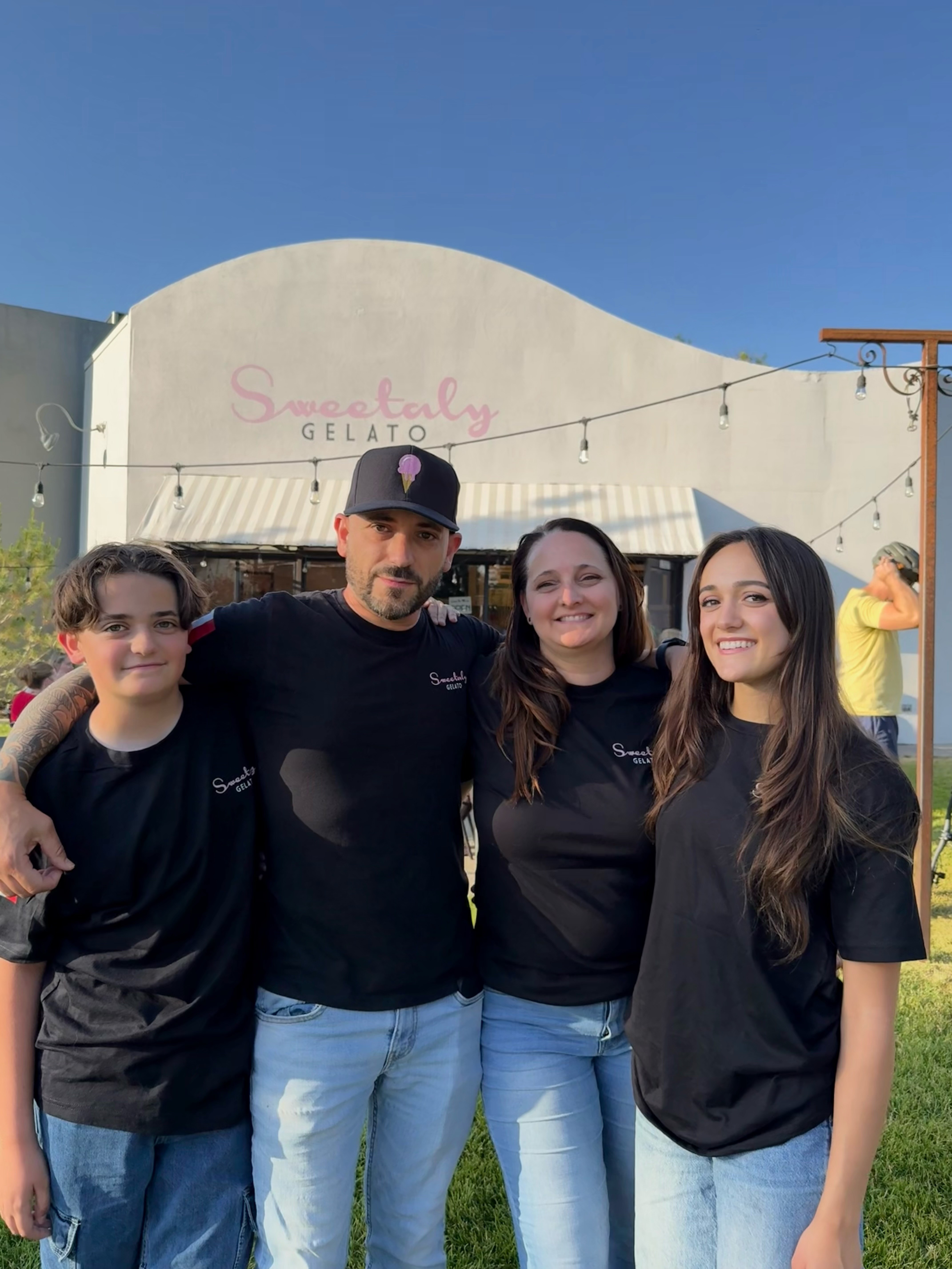 Four people standing outdoors in front of a building with a sign that reads "Sweetably Gelato." They are smiling and wearing black shirts.