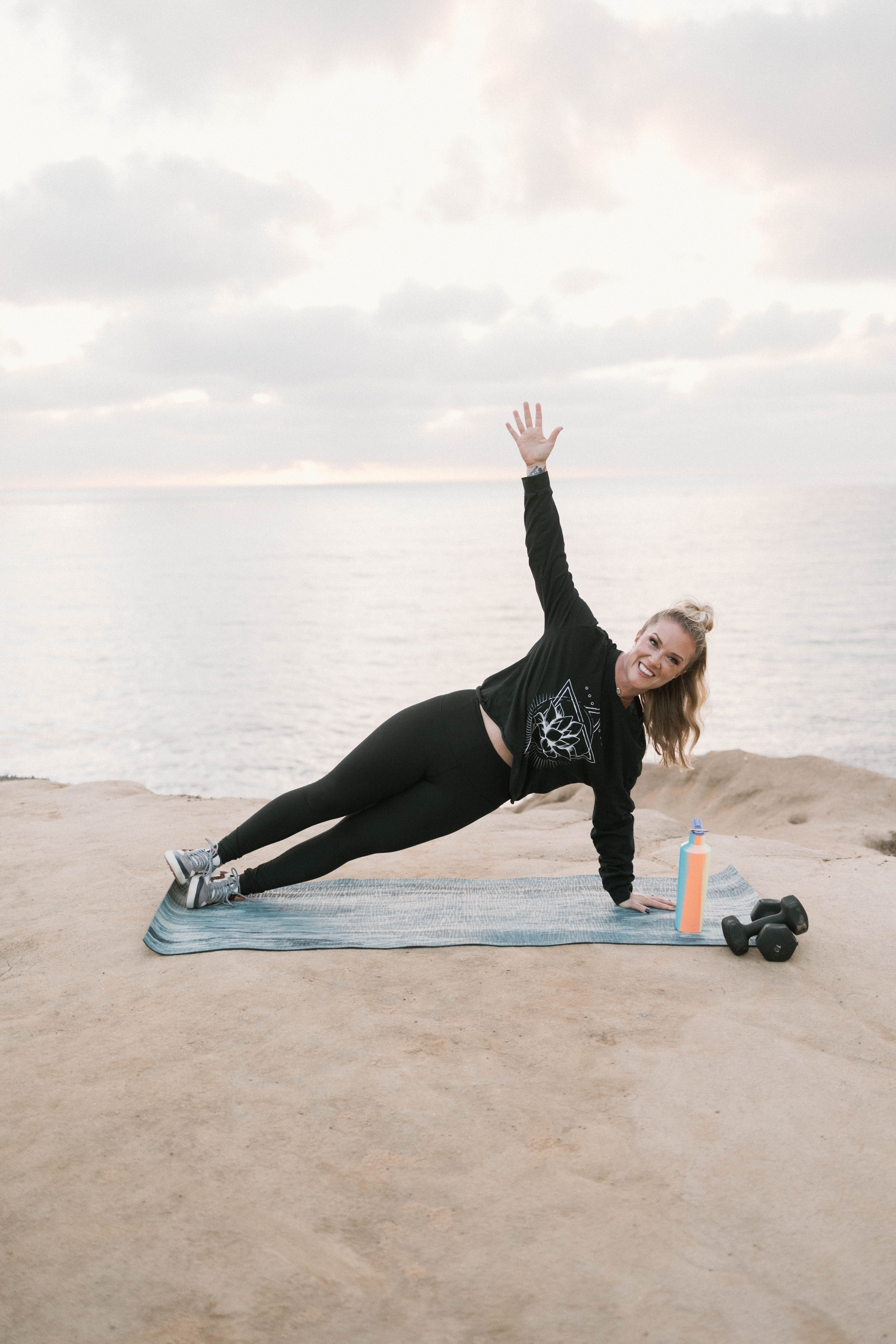 Woman exercising outdoors on a mat near water, raising one arm, with water and cloudy sky in background.