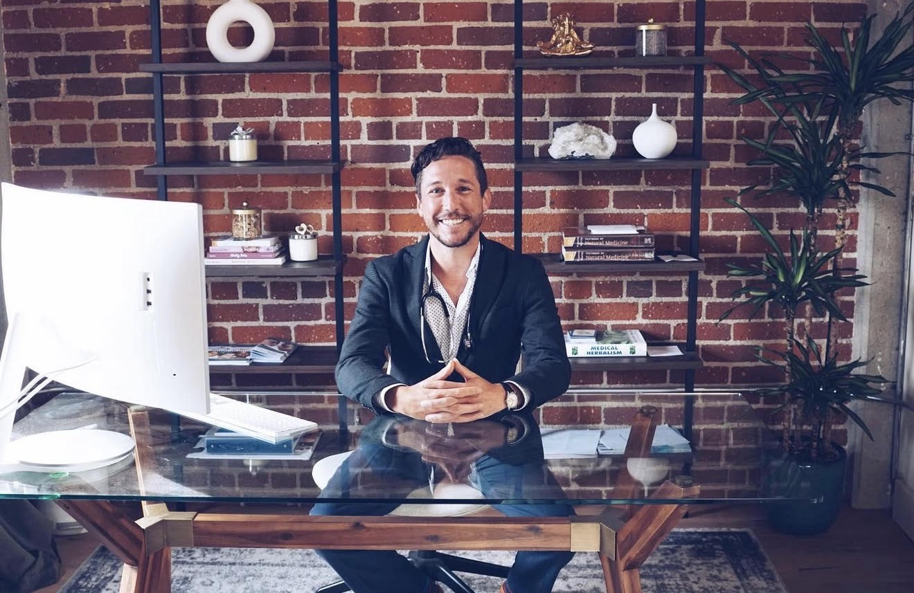 Man in suit sitting at glass desk with computer, brick wall and shelves behind him, smiling.