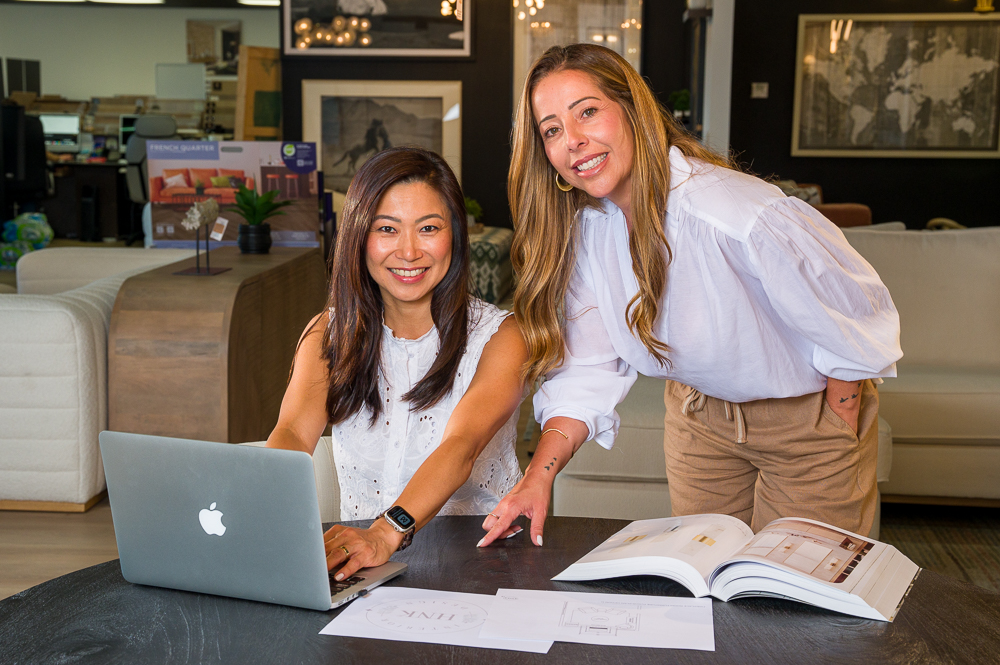 Two women working together at a table with a laptop and open books in a modern office or lounge area.