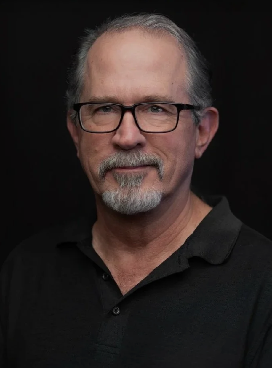 Portrait of a middle-aged man with glasses, gray hair, and a beard, wearing a black polo shirt, against a dark background.