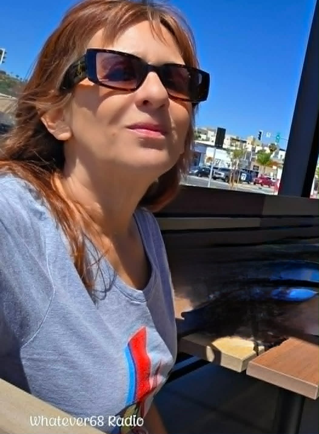 Woman with sunglasses sitting outdoors at a table, with buildings and cars in the background.