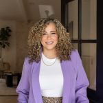 Smiling woman with curly hair wearing a purple blazer and white top, standing indoors near a black door frame.