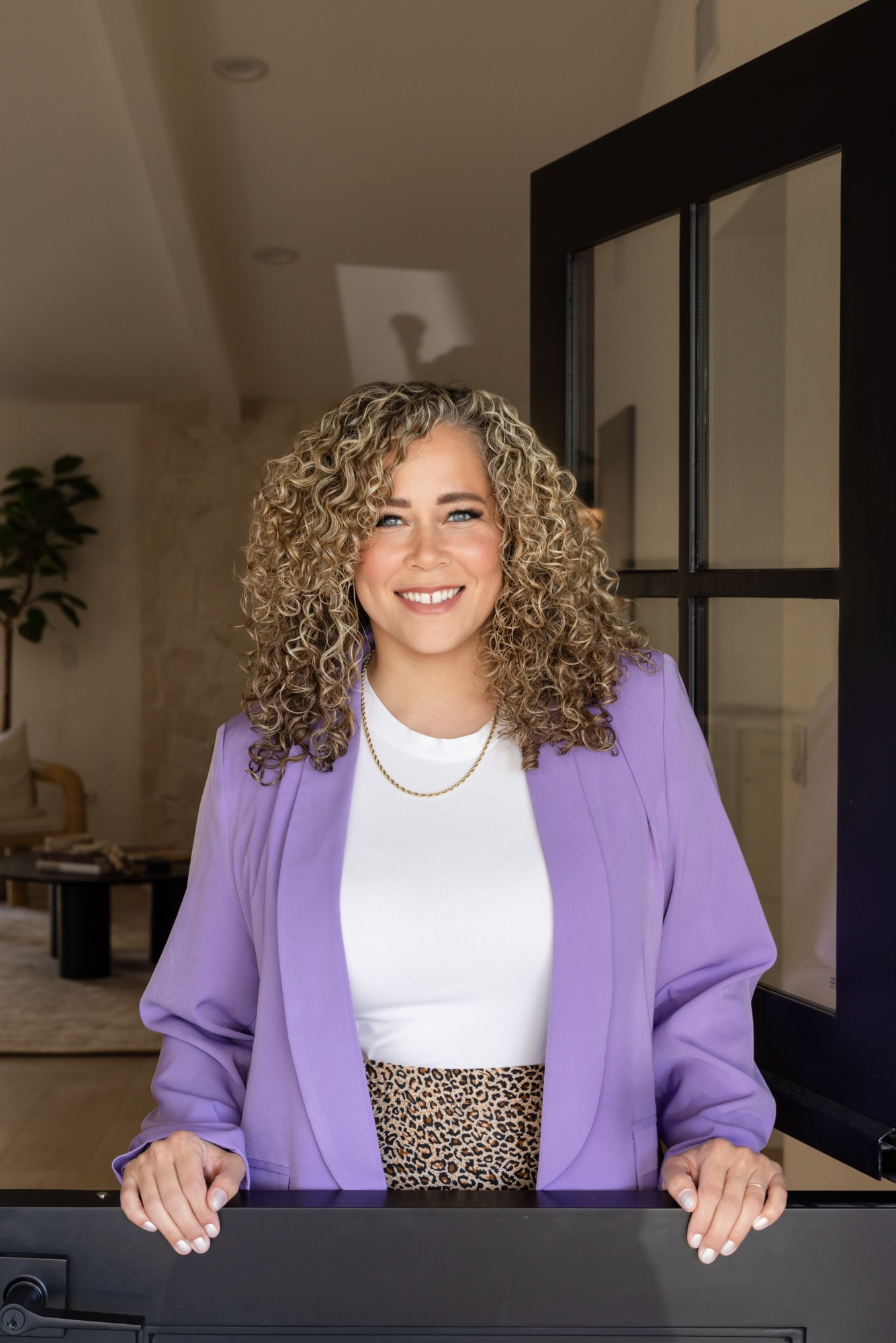 Smiling woman with curly hair wearing a purple blazer and white top, standing indoors near a black door frame.