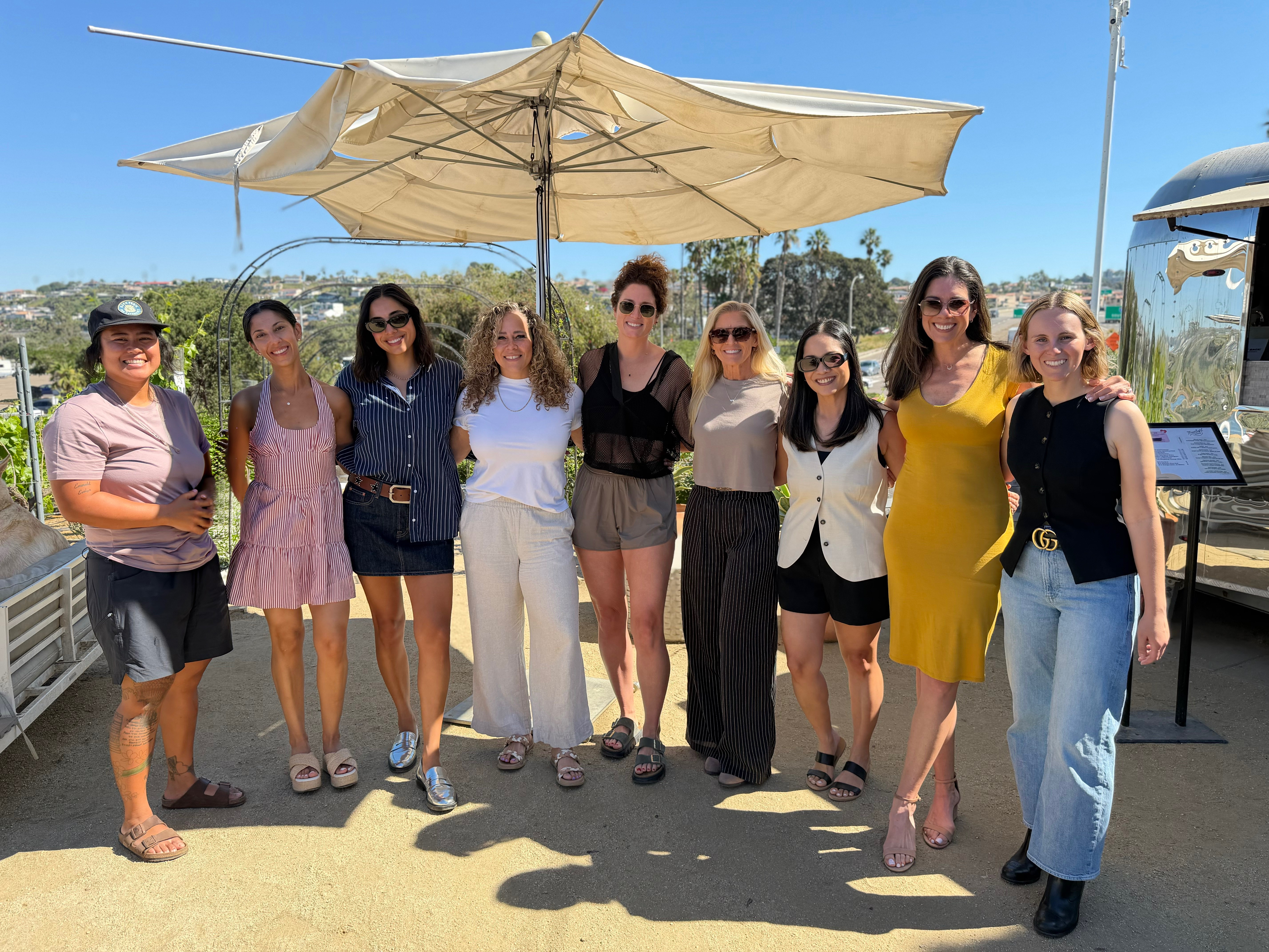 Group of nine women standing outdoors under a large umbrella, smiling, with a boat and blue sky in background.