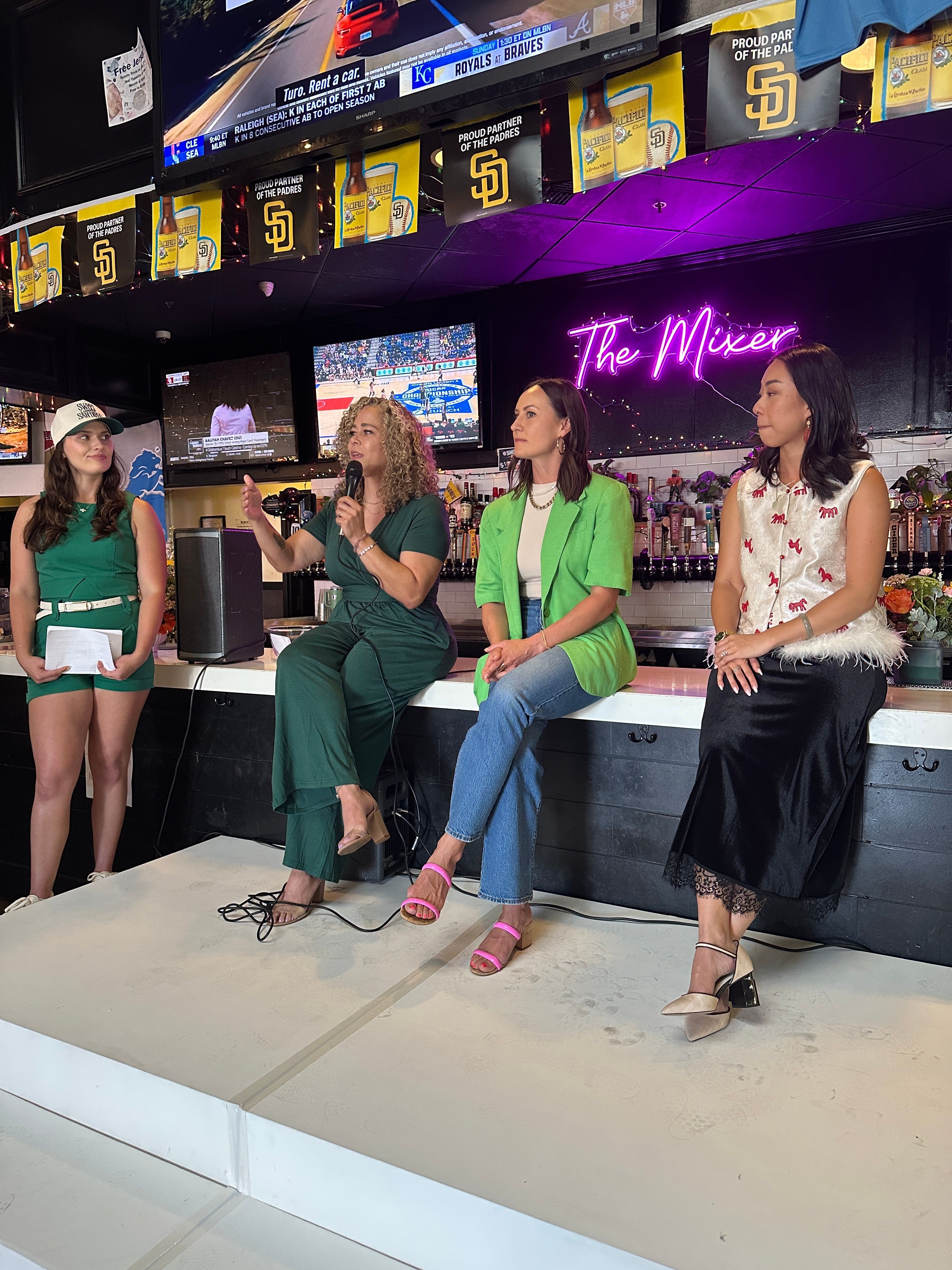 Four women stand together in a store, posing for a photo, with shelves and a purple-lit sign in the background.