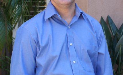 Smiling man in a blue shirt standing outdoors with plants in background.