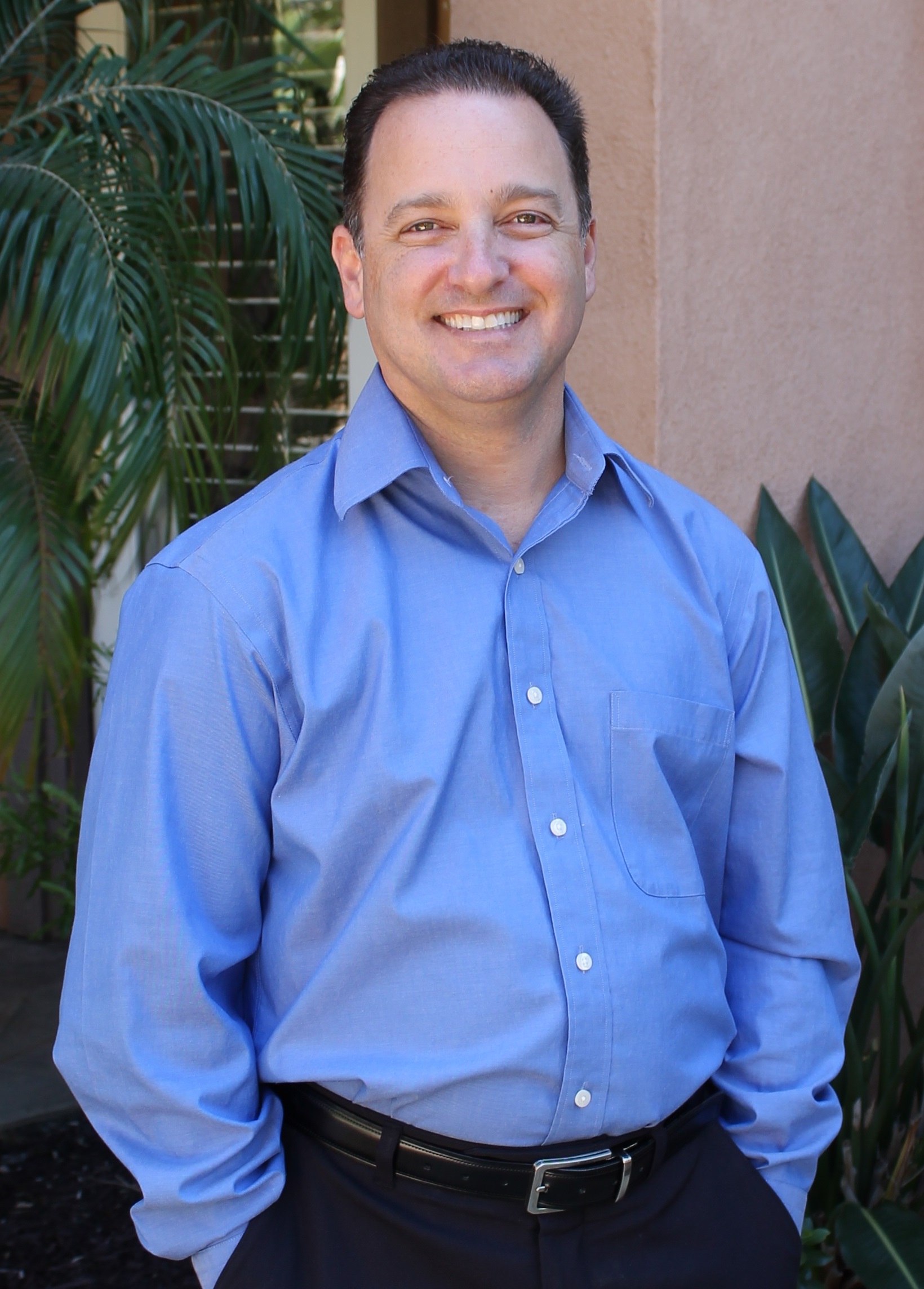 Smiling man in a blue shirt standing outdoors with plants in background.