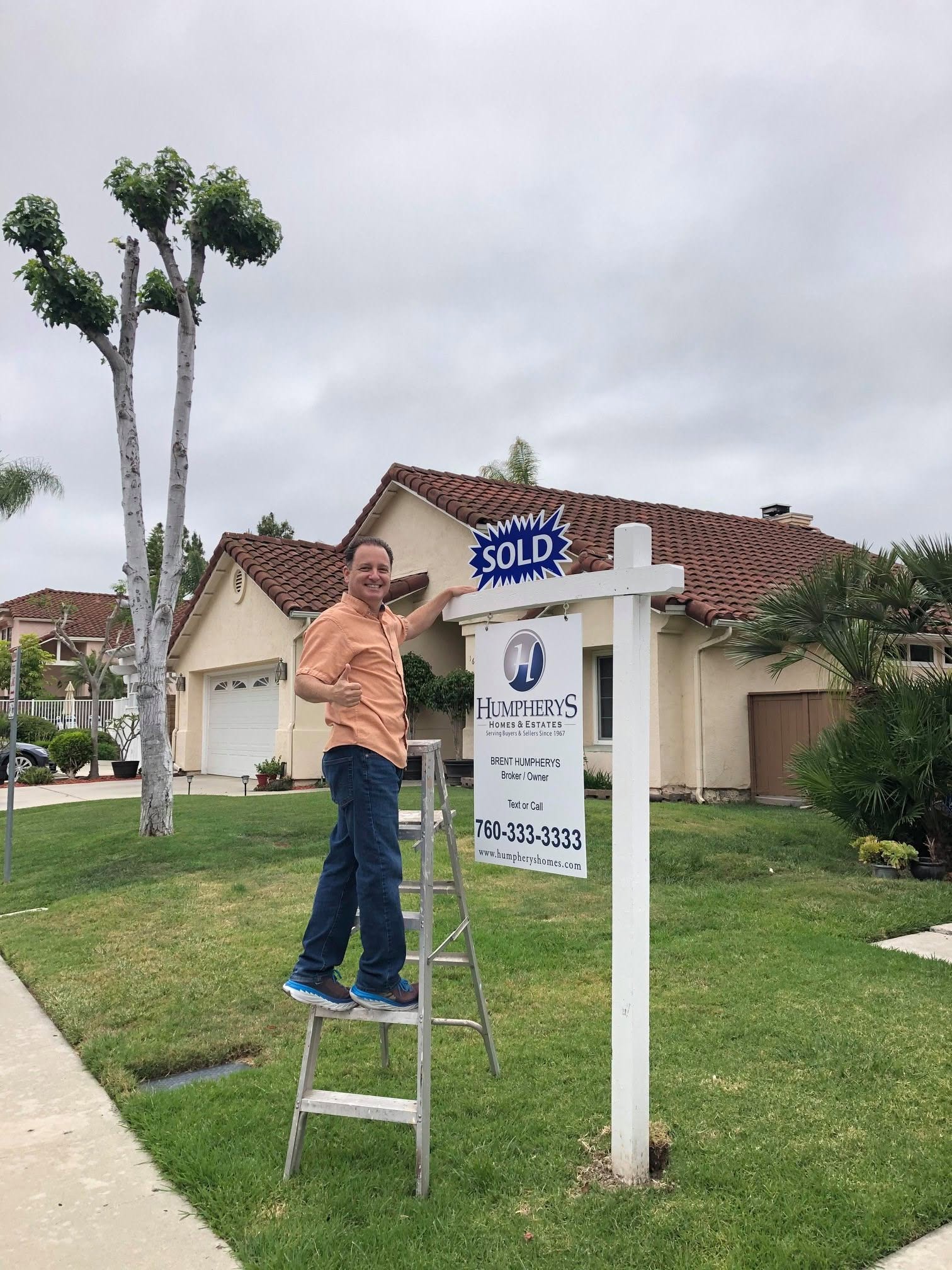 Person standing on a ladder next to a sold sign in front of a house, with trees and a cloudy sky in the background.