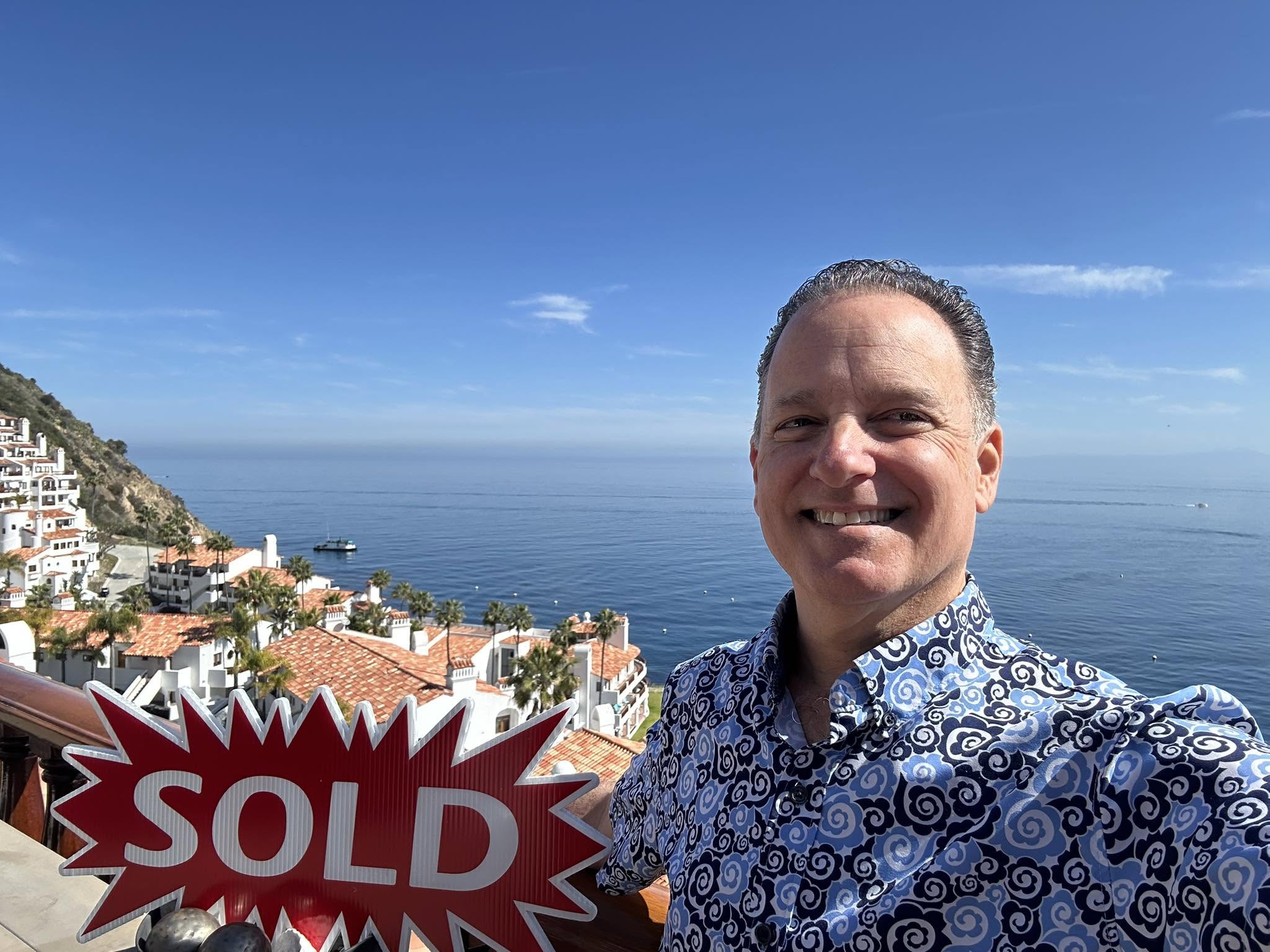 Smiling man taking selfie with coastal town and ocean in background under blue sky