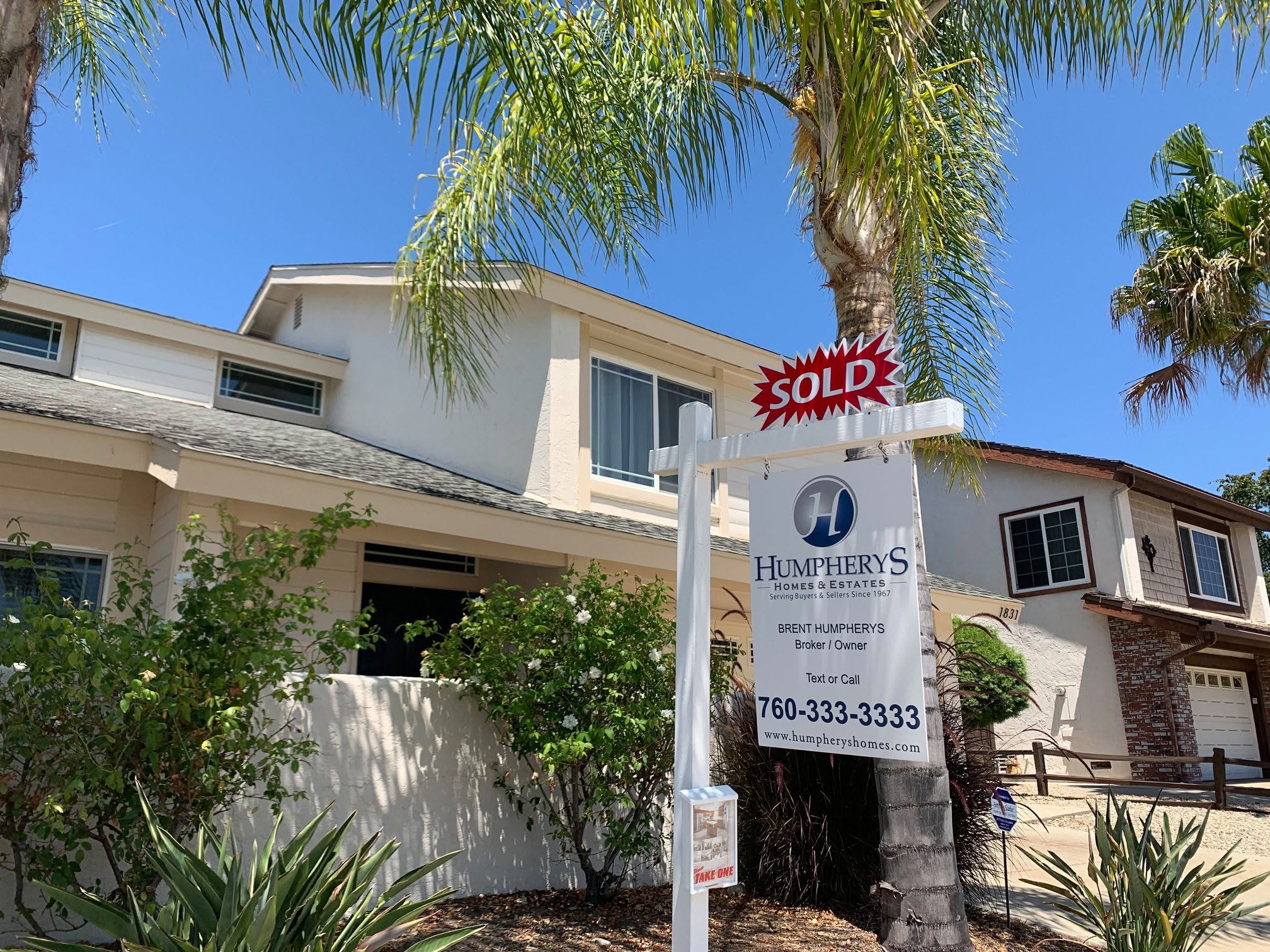 House with a 'Sold' sign on a palm tree, blue sky background, lush greenery, neighboring house visible.