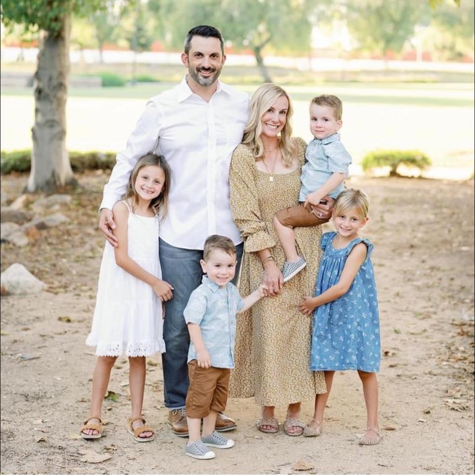 Family of six outdoors, smiling, standing on dirt ground with trees and park in background.