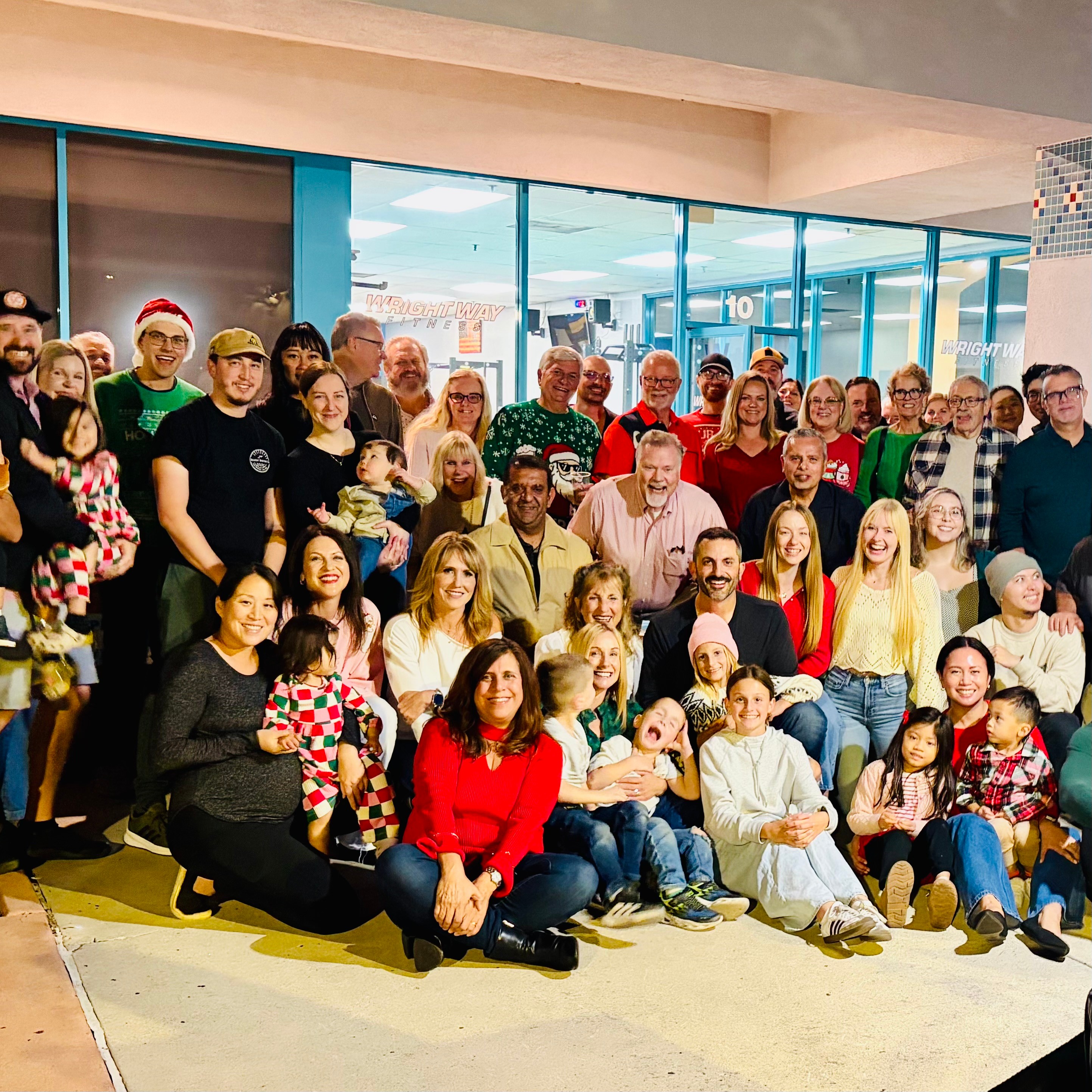 Group of people, including children, gathered indoors for a photo, some holding gifts, with a glass window background.