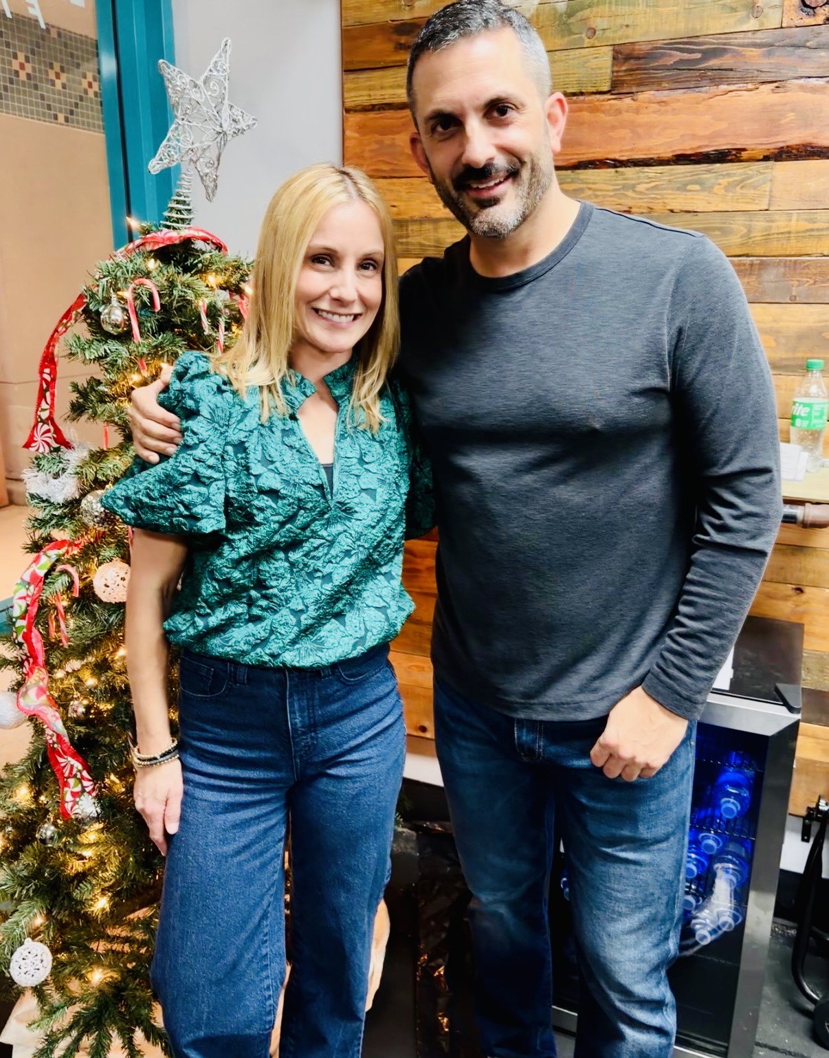 A smiling woman and man stand indoors near a decorated Christmas tree, with a wooden wall behind them.