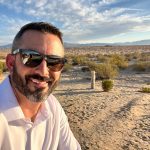 Man with sunglasses and beard taking selfie outdoors on sandy terrain with blue sky and clouds.