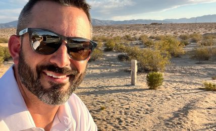 Man with sunglasses and beard taking selfie outdoors on sandy terrain with blue sky and clouds.