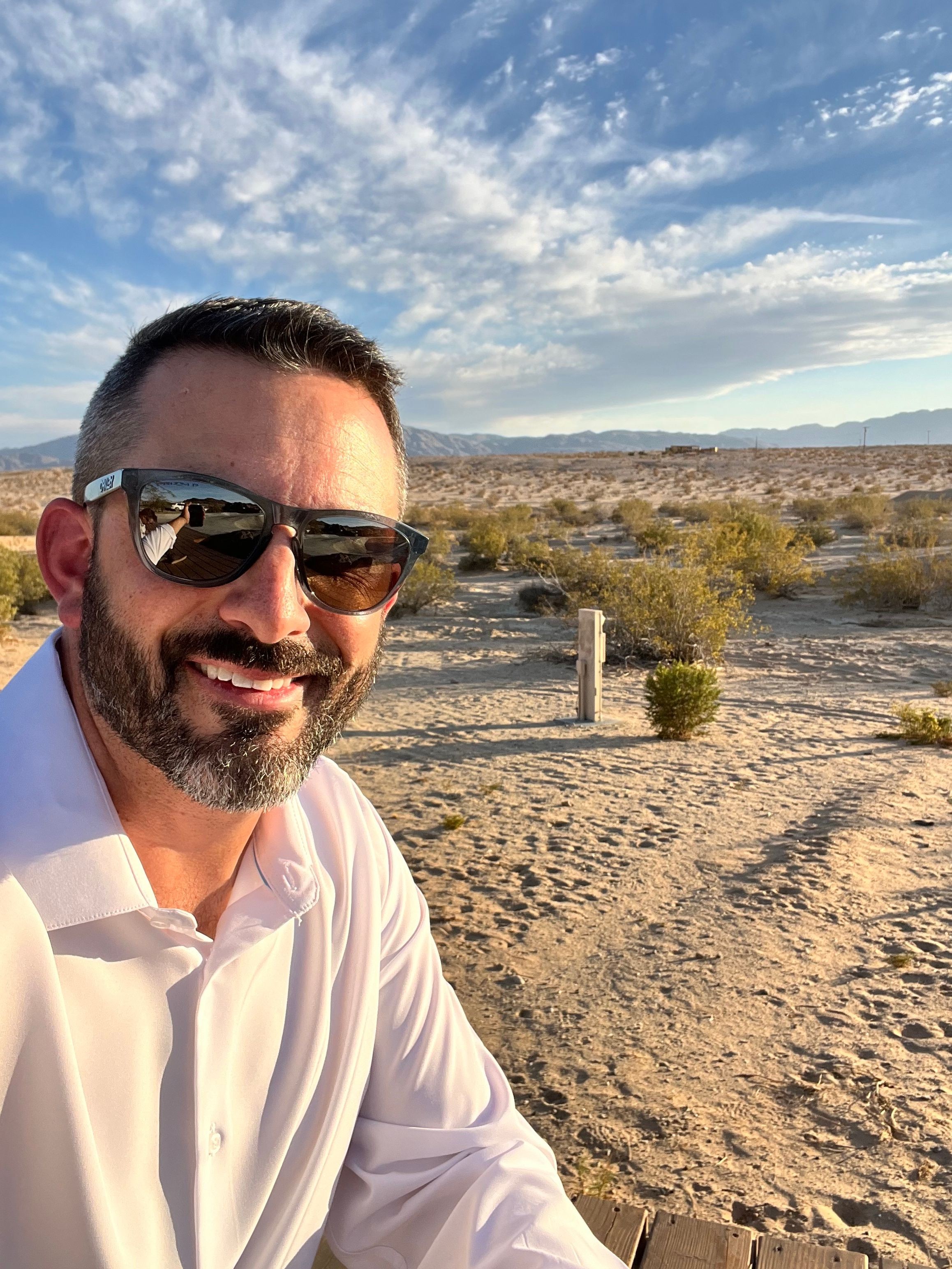 Man with sunglasses and beard taking selfie outdoors on sandy terrain with blue sky and clouds.