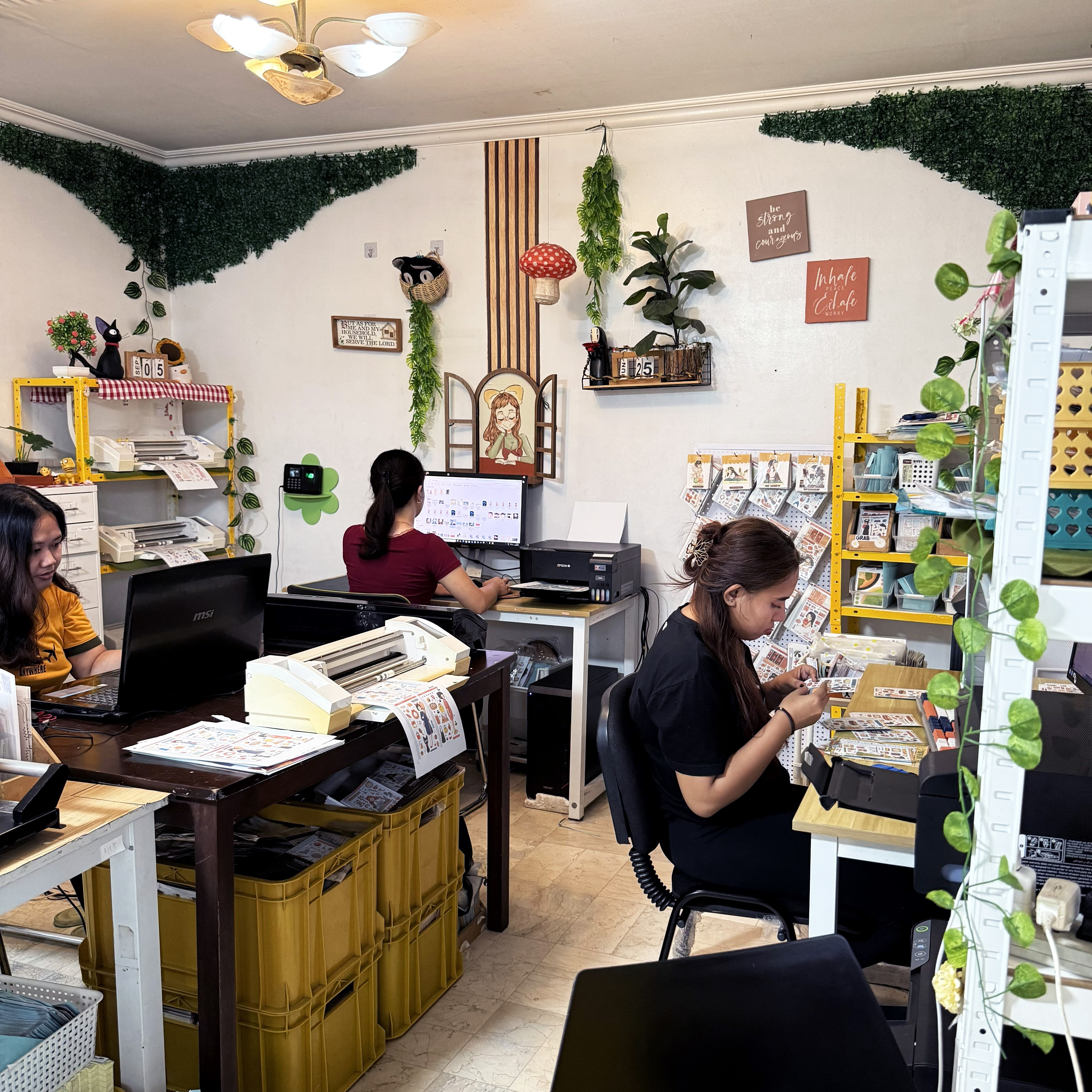 People working at desks in a small office with shelves, plants, and decorations on the walls.