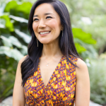 Smiling woman with long dark hair in a colorful sleeveless dress outdoors, greenery in background.