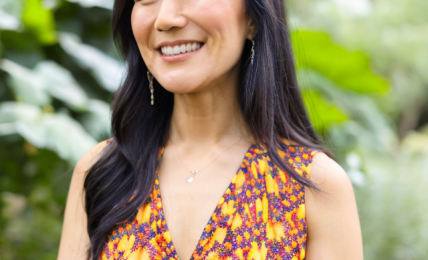 Smiling woman with long dark hair in a colorful sleeveless dress outdoors, greenery in background.