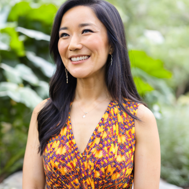 Smiling woman with long dark hair in a colorful sleeveless dress outdoors, greenery in background.