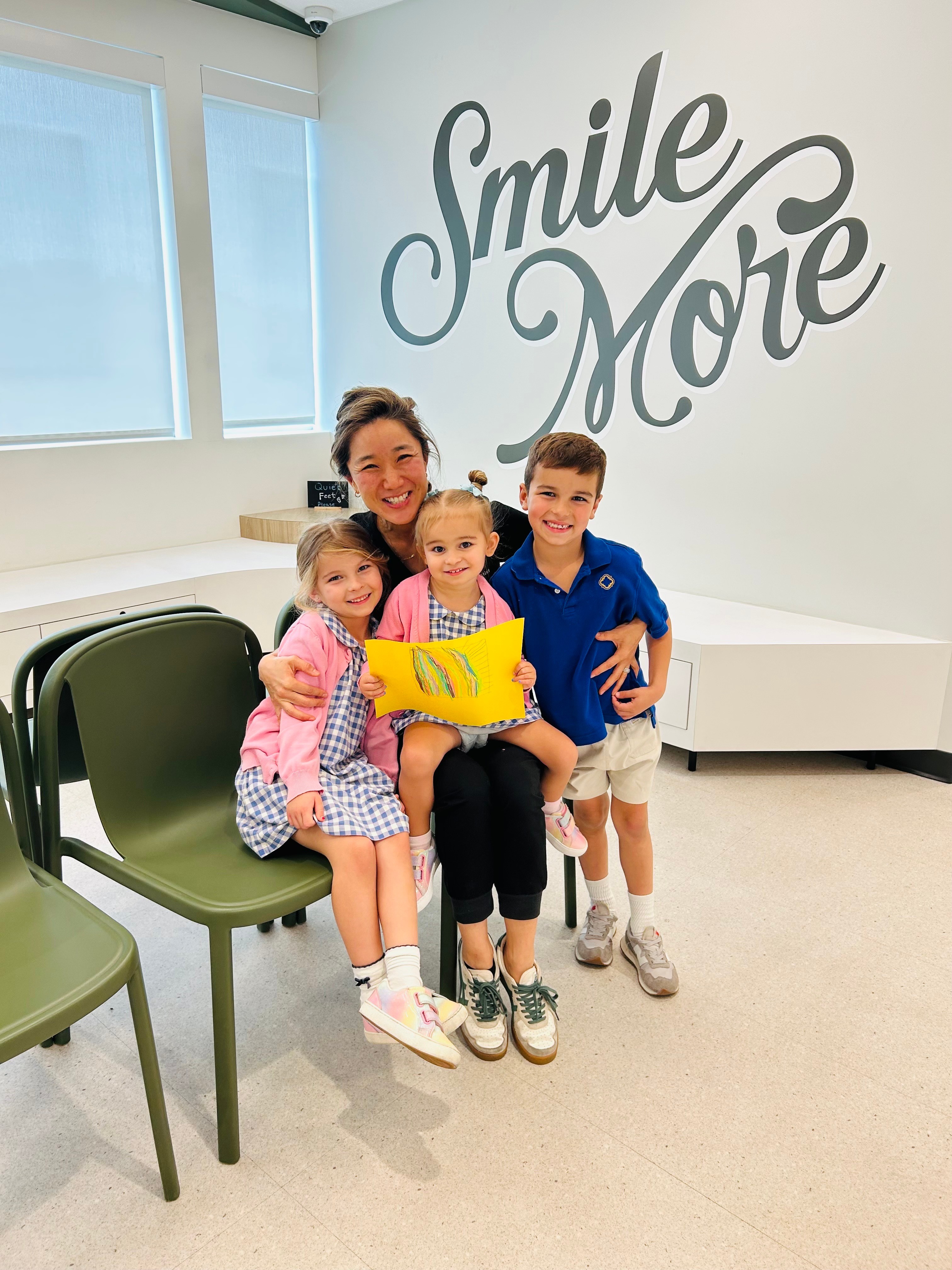 A woman and three children sitting on chairs in a room with a wall reading 'Smile More'.