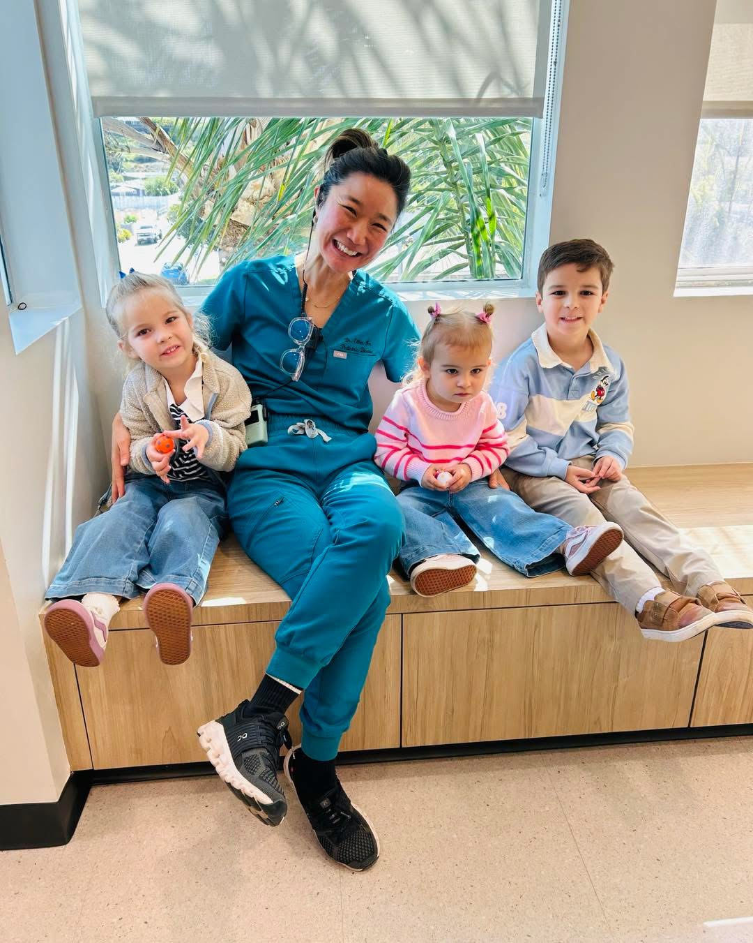 A woman in blue scrubs sitting on a bench with three children, smiling, in a bright room with large windows and greenery outside.
