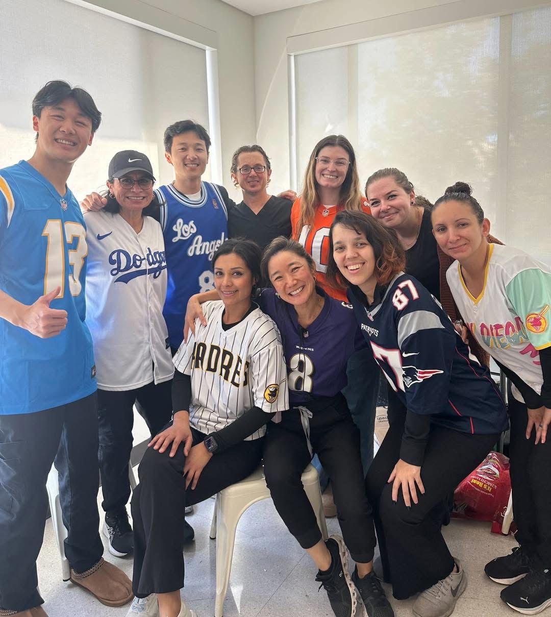 Group of ten young women and men smiling indoors, some wearing sports jerseys, in front of large windows.