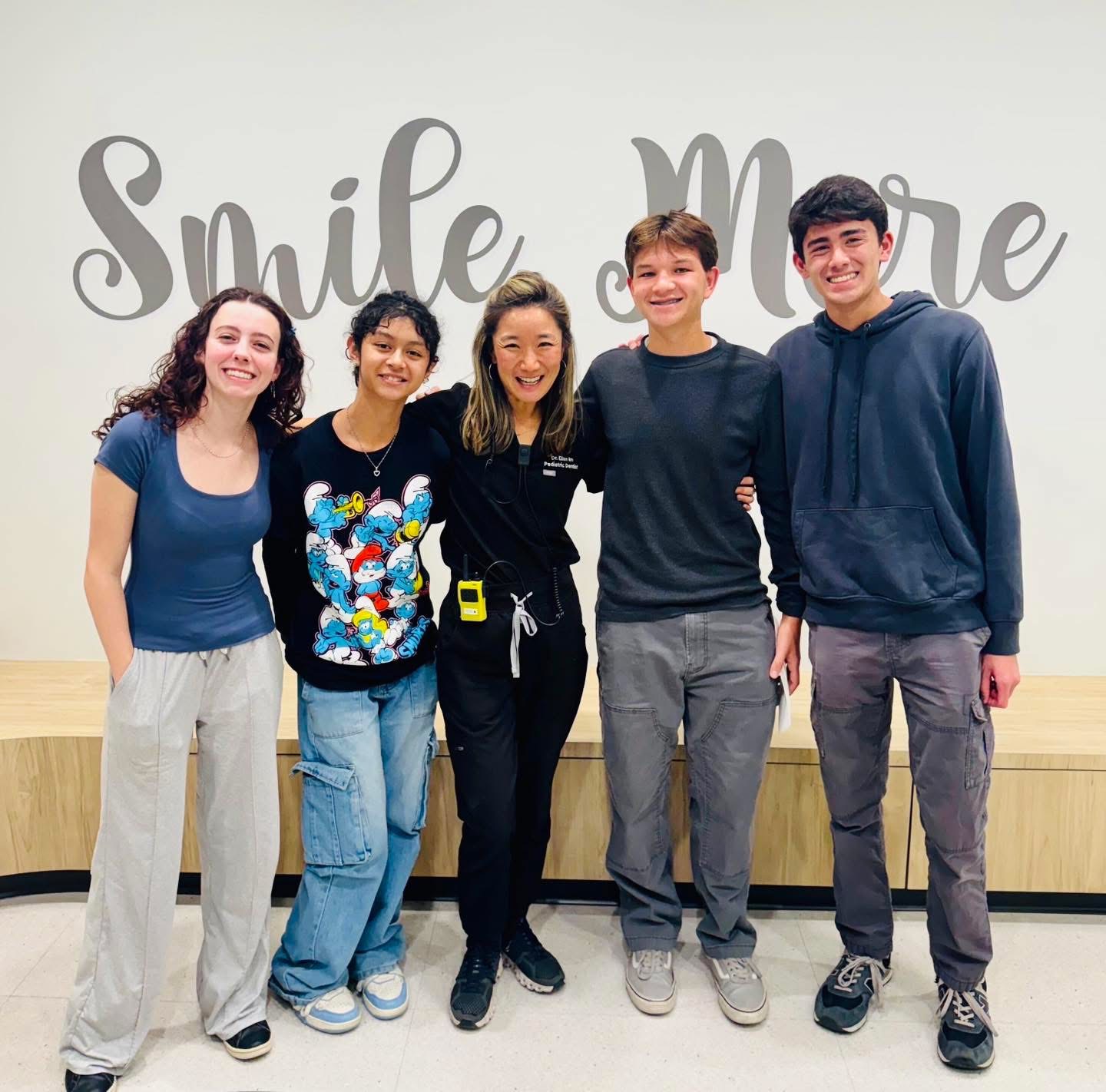 Five people standing together in front of a wall with the words 'Smile More' written on it, smiling and posing for the photo.