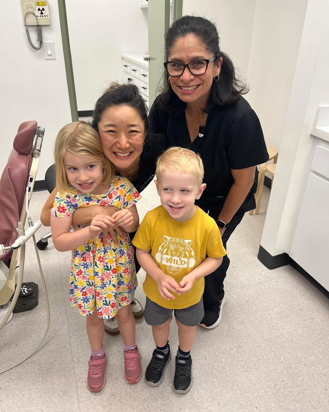 Two children and two women smiling indoors, one woman wearing glasses, children holding hands, cheerful atmosphere.