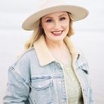 Smiling woman wearing a wide-brimmed hat and denim jacket outdoors, light background.