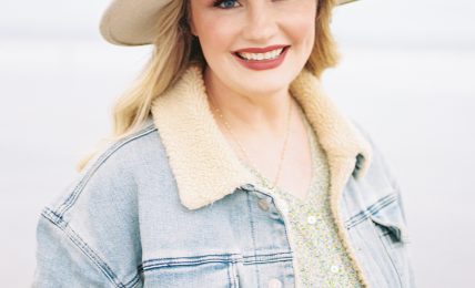 Smiling woman wearing a wide-brimmed hat and denim jacket outdoors, light background.