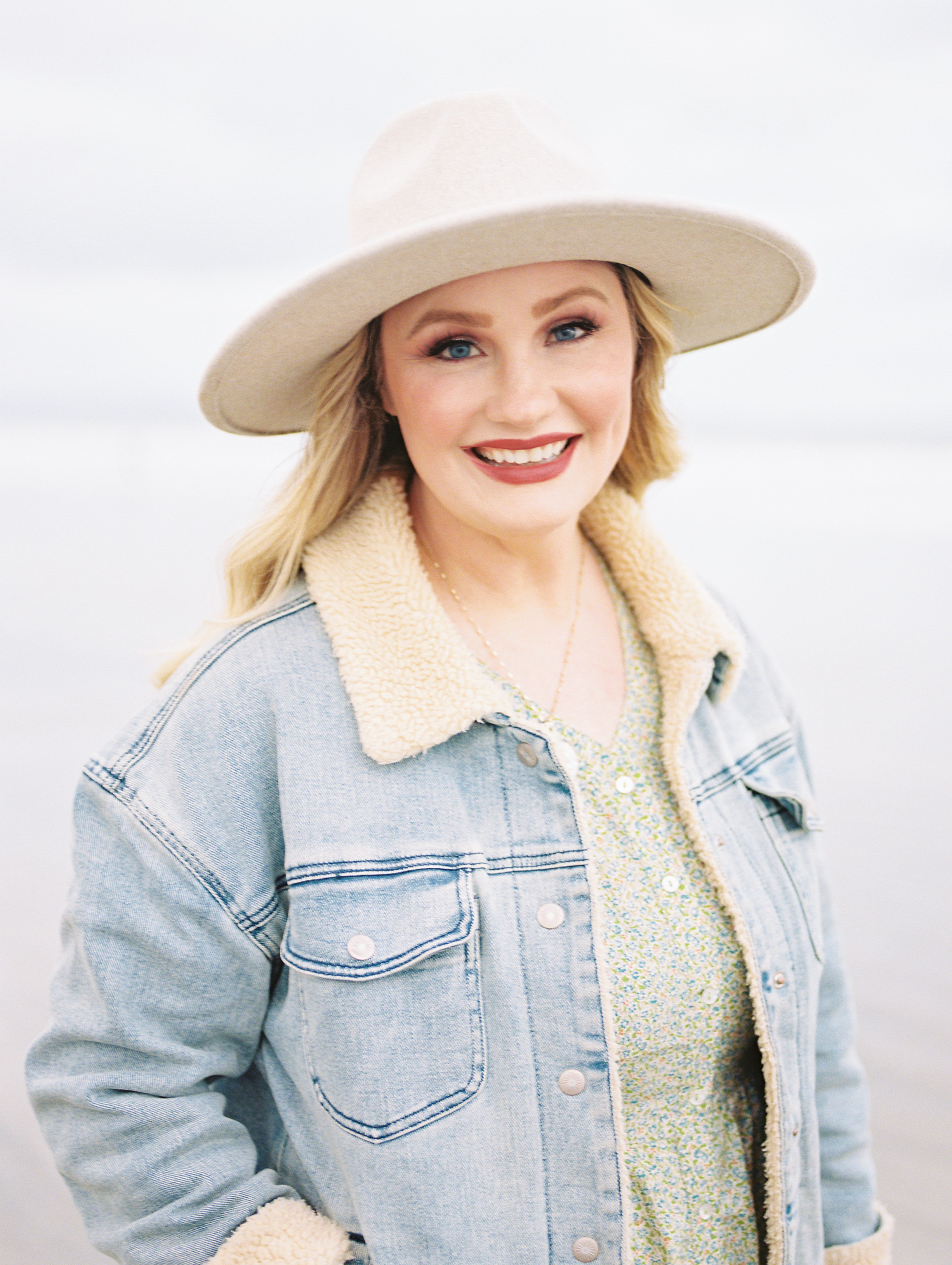 Smiling woman wearing a wide-brimmed hat and denim jacket outdoors, light background.