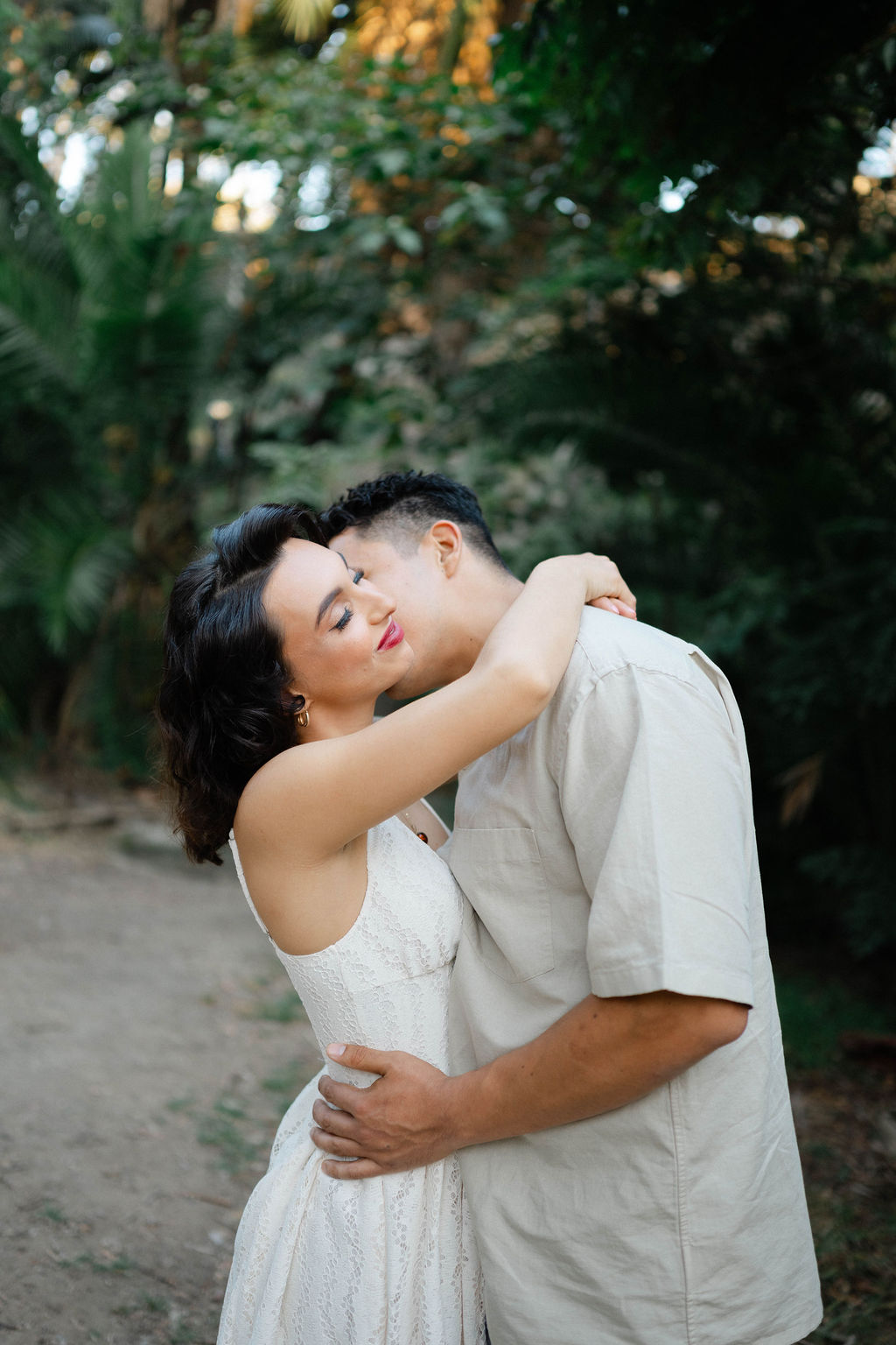 A couple embraces outdoors with trees in the background, woman with dark hair and white dress, man in light shirt.