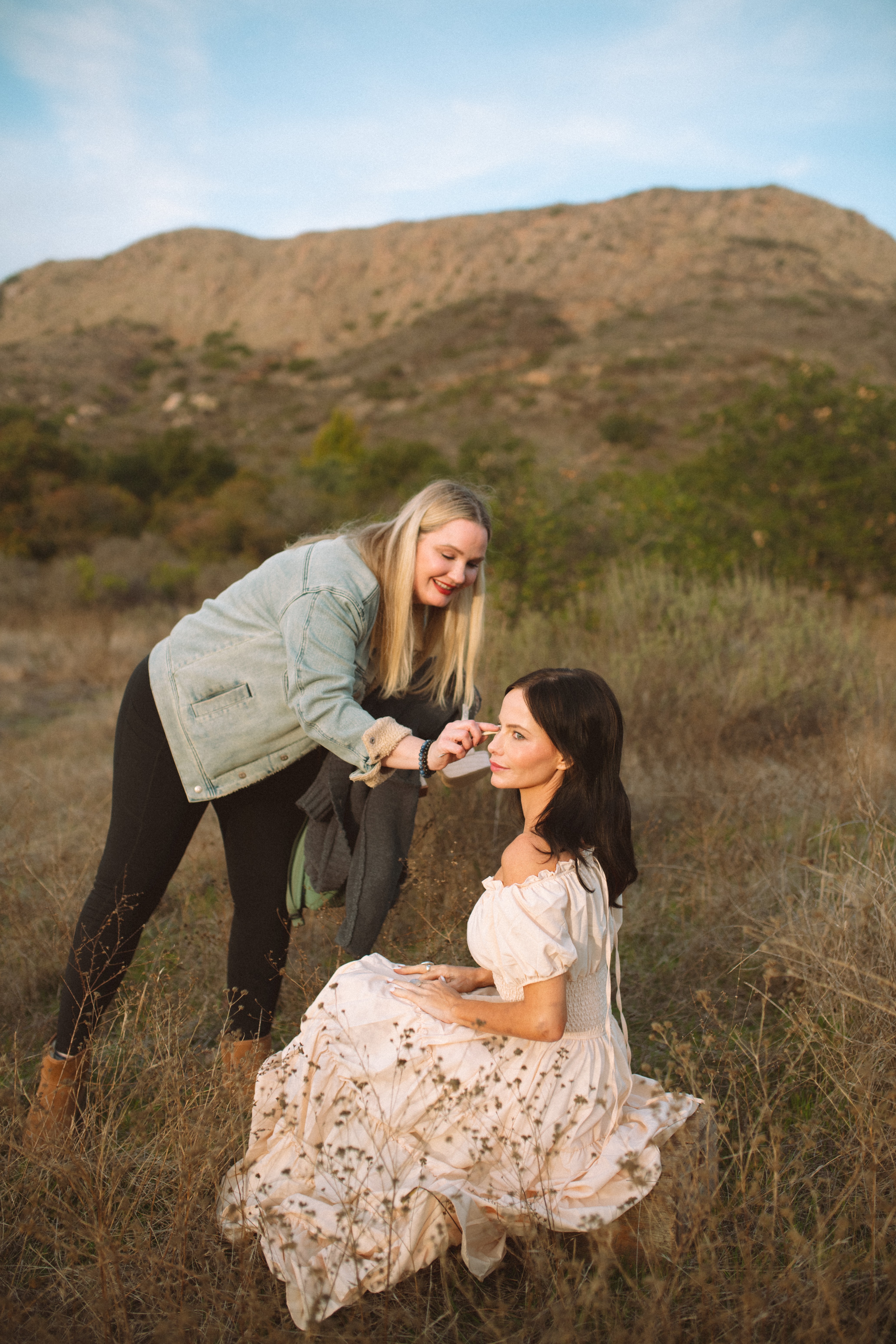 Two women outdoors, one standing and applying makeup to the other sitting on grass, with hills in background.