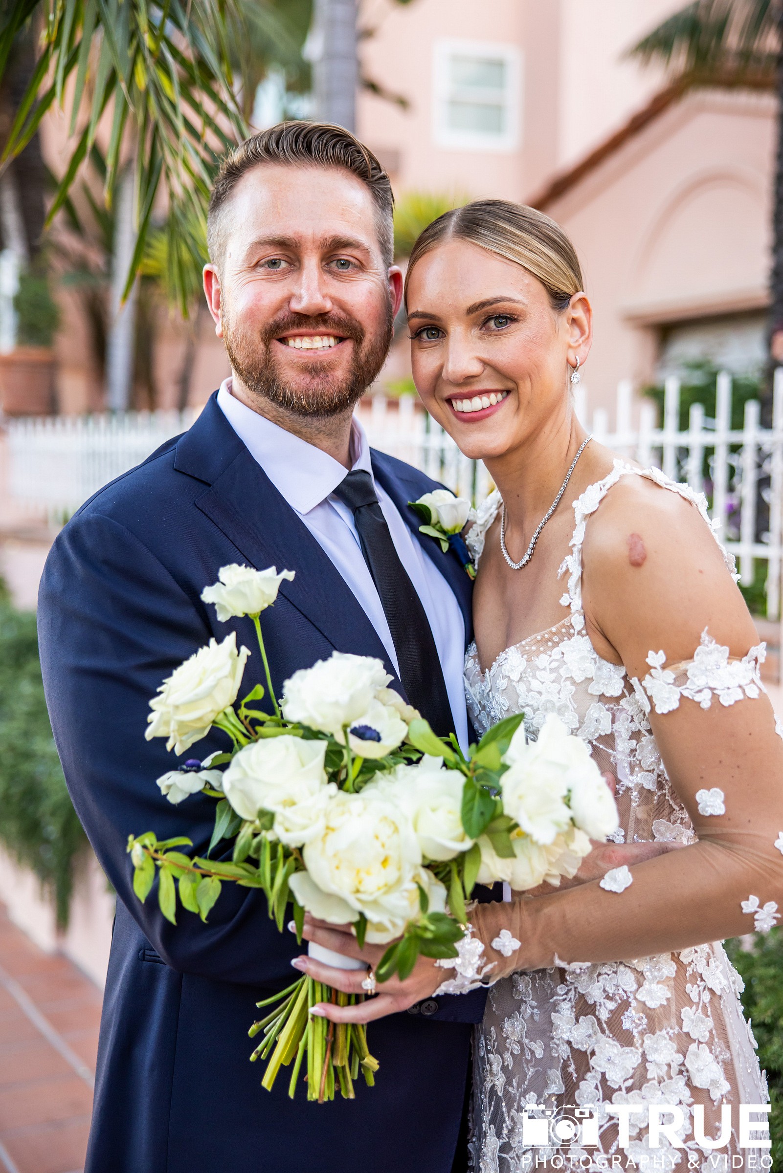 A smiling couple in wedding attire holding a bouquet of white flowers outdoors, with greenery and a building in the background.