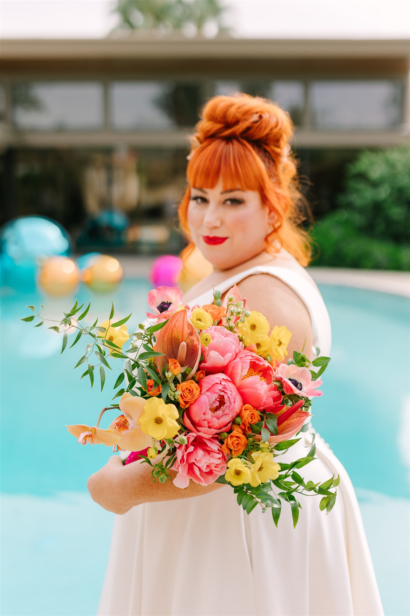Woman with red hair in a white dress holding a colorful bouquet near a pool, background with trees and pool floats.