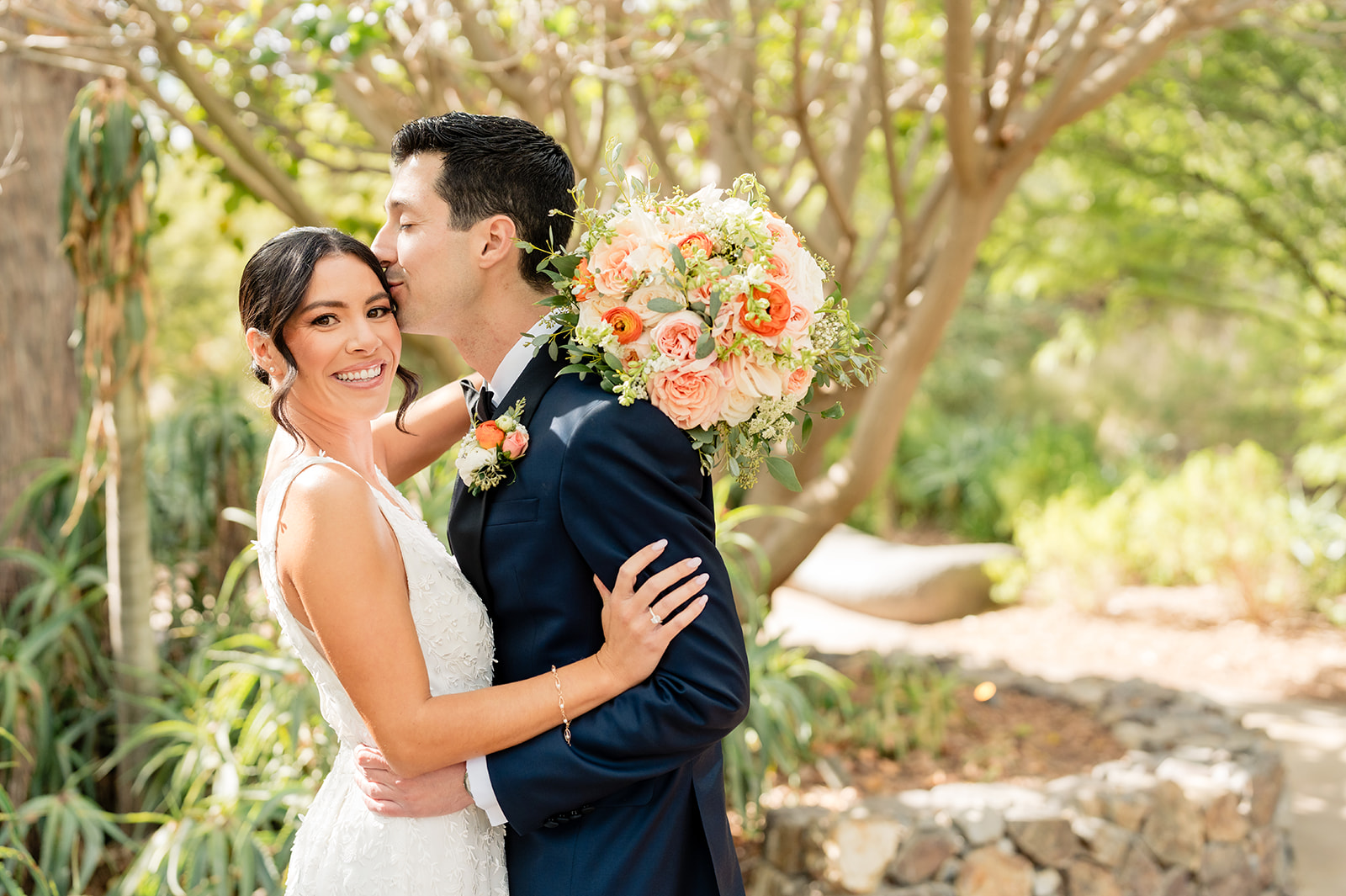 A bride and groom outdoors, smiling, with trees and greenery in the background.