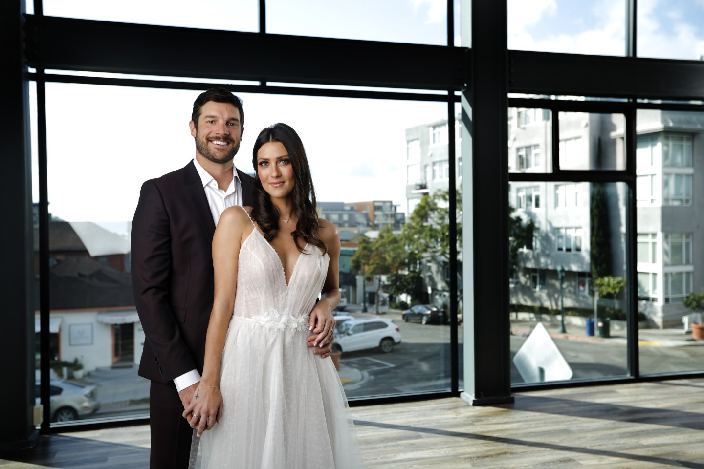 A smiling man and woman in wedding attire standing inside a modern building with large windows, cityscape outside.