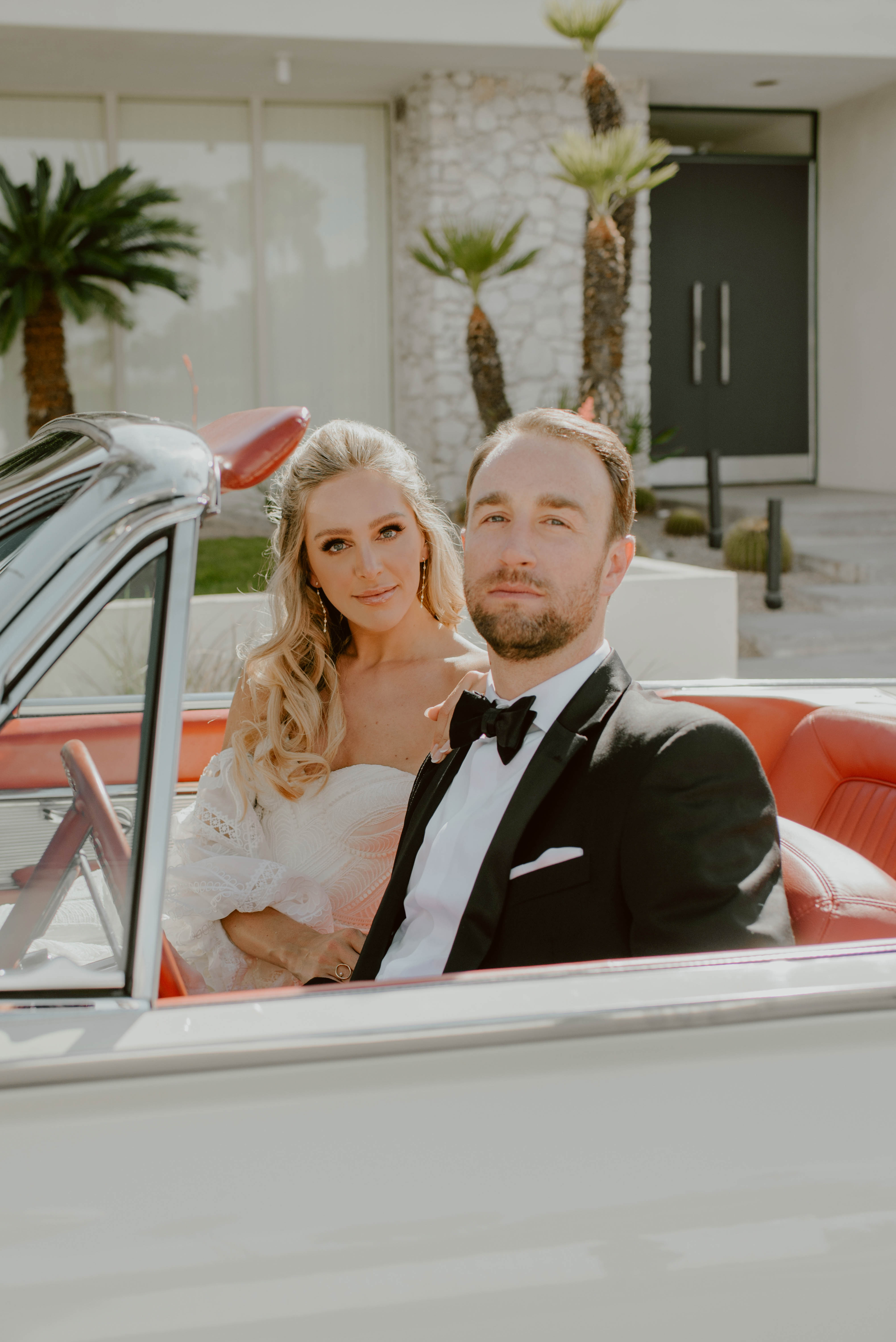 A couple in wedding attire sitting in a vintage car outdoors, with palm trees and modern building in background.