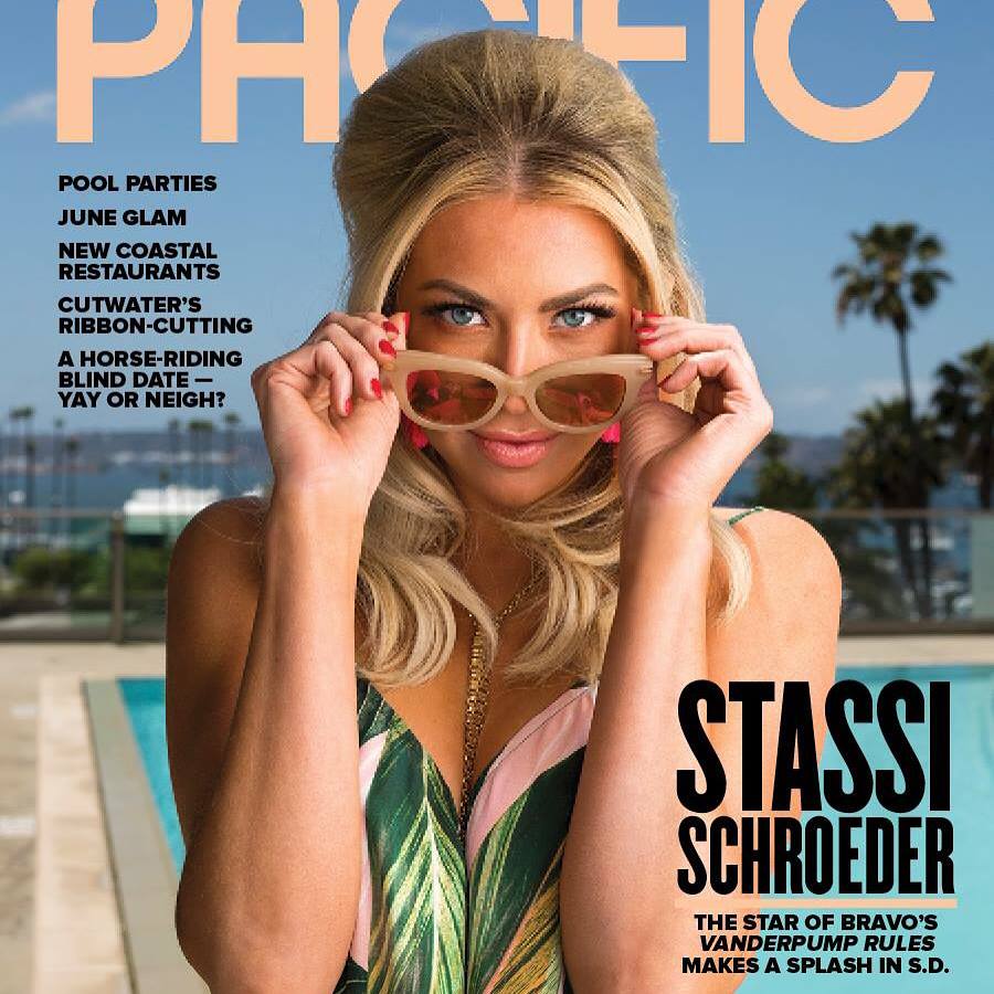 Woman with blonde hair holding sunglasses, smiling, outdoors with pool and ocean in background.