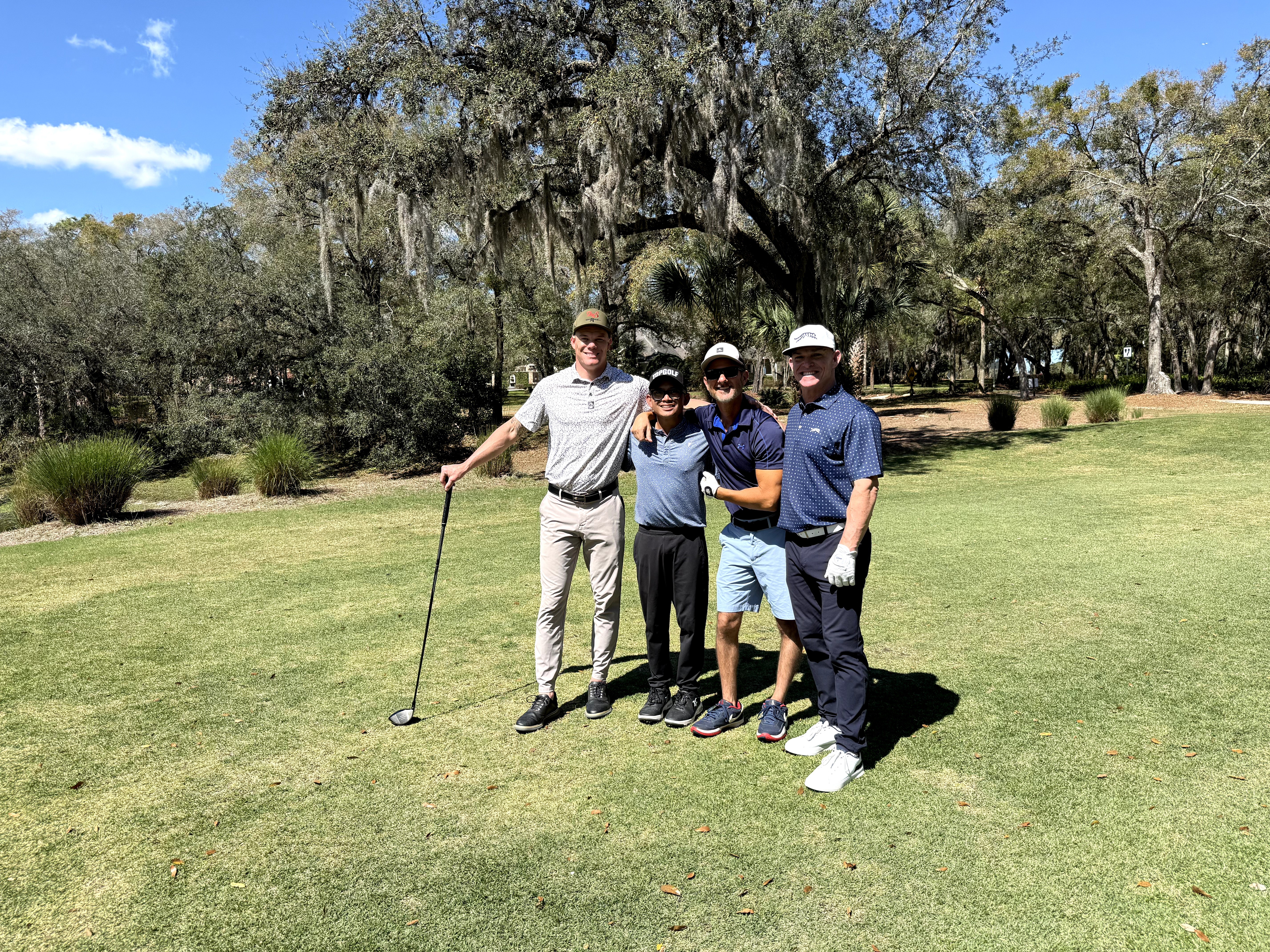 Four people standing on a golf course with trees and grass, posing for a photo, one holding a golf club.