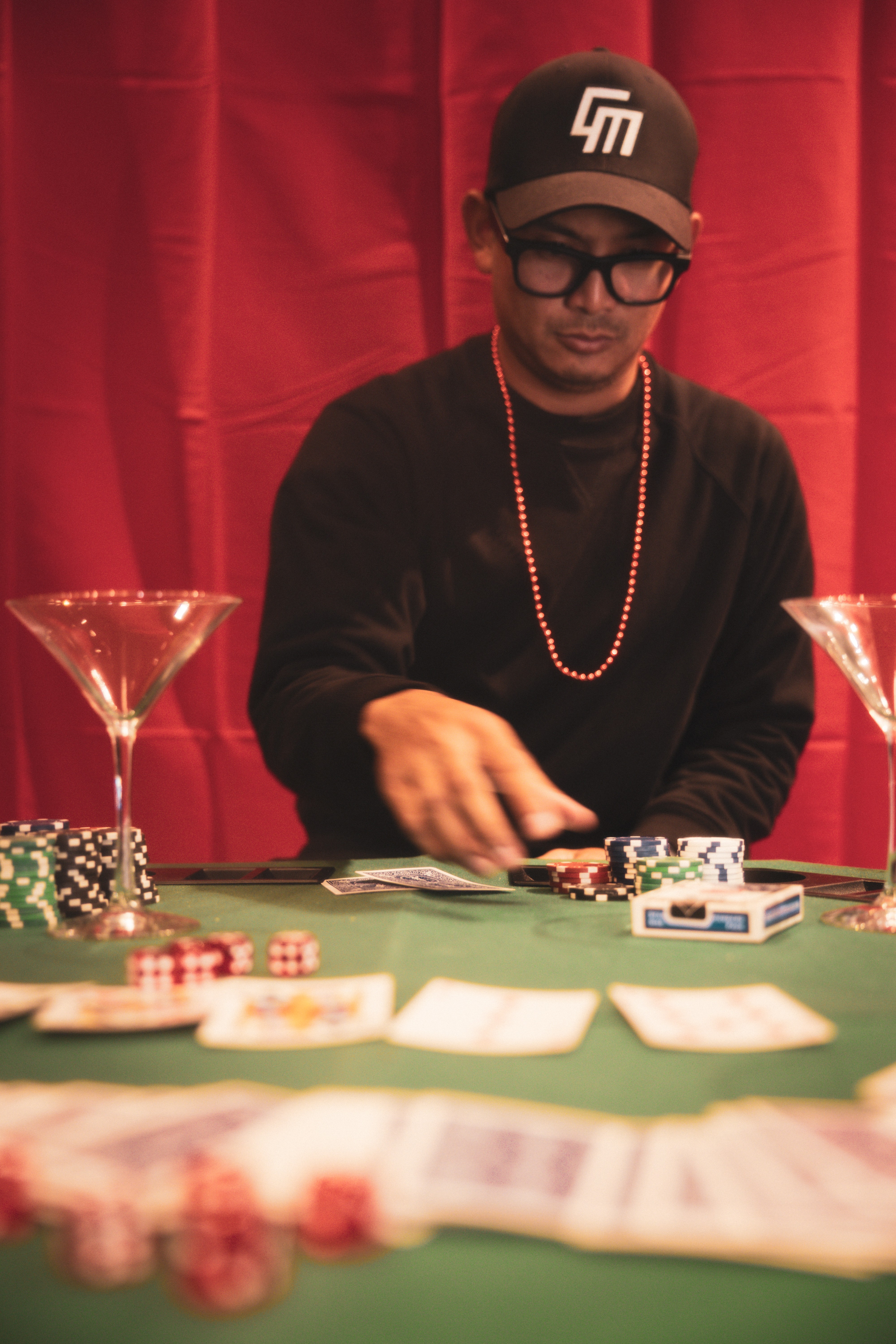 Person wearing glasses, a cap, and a beaded necklace, playing poker at a table with chips and cards, red curtain background.