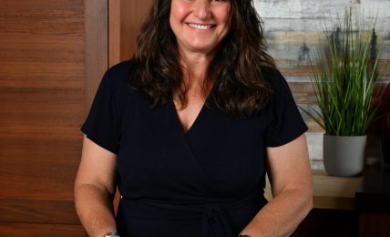 Smiling woman with dark hair in a black top holding papers, standing at a marble table in a room with wooden and plant decor.