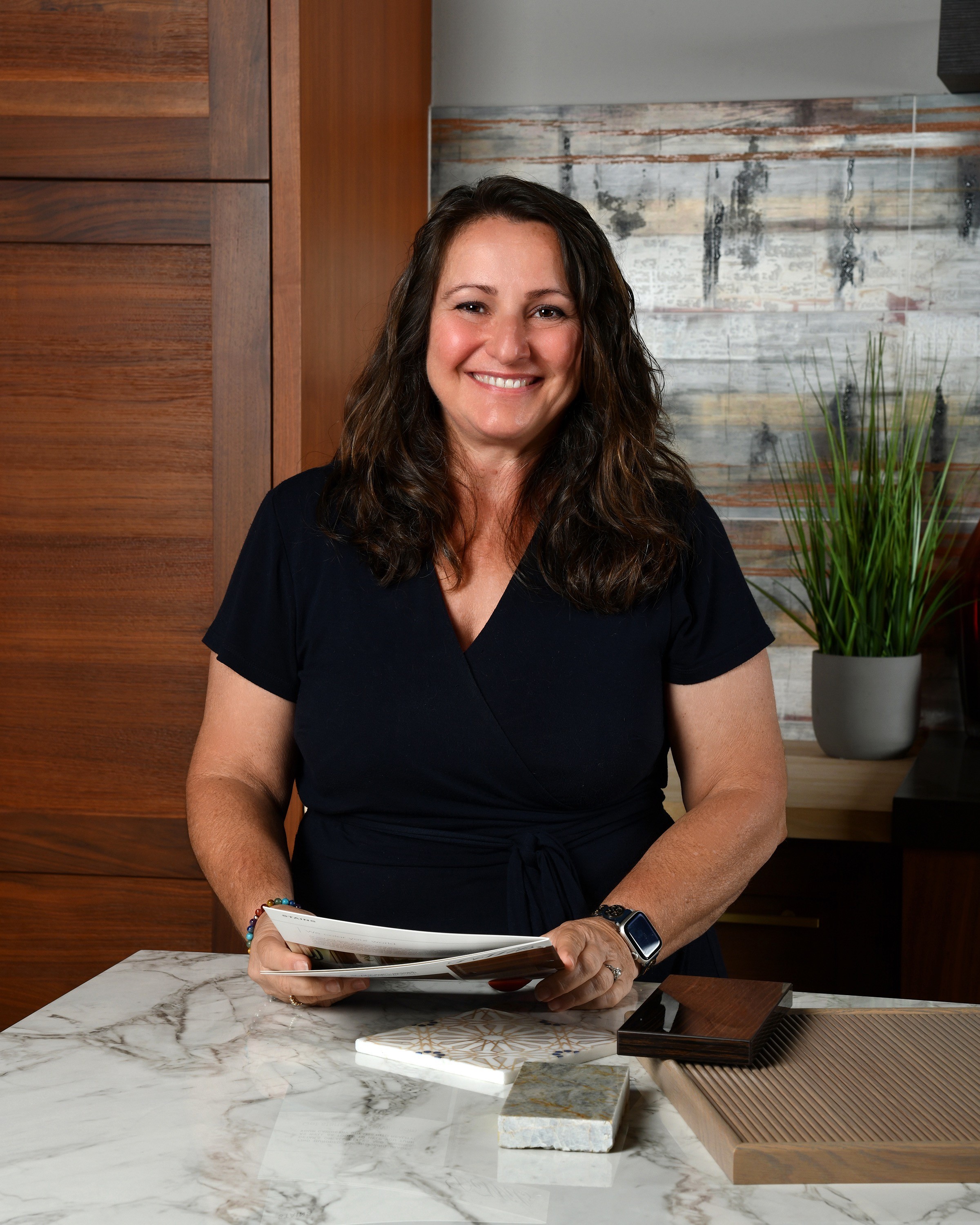 Smiling woman with dark hair in a black top holding papers, standing at a marble table in a room with wooden and plant decor.