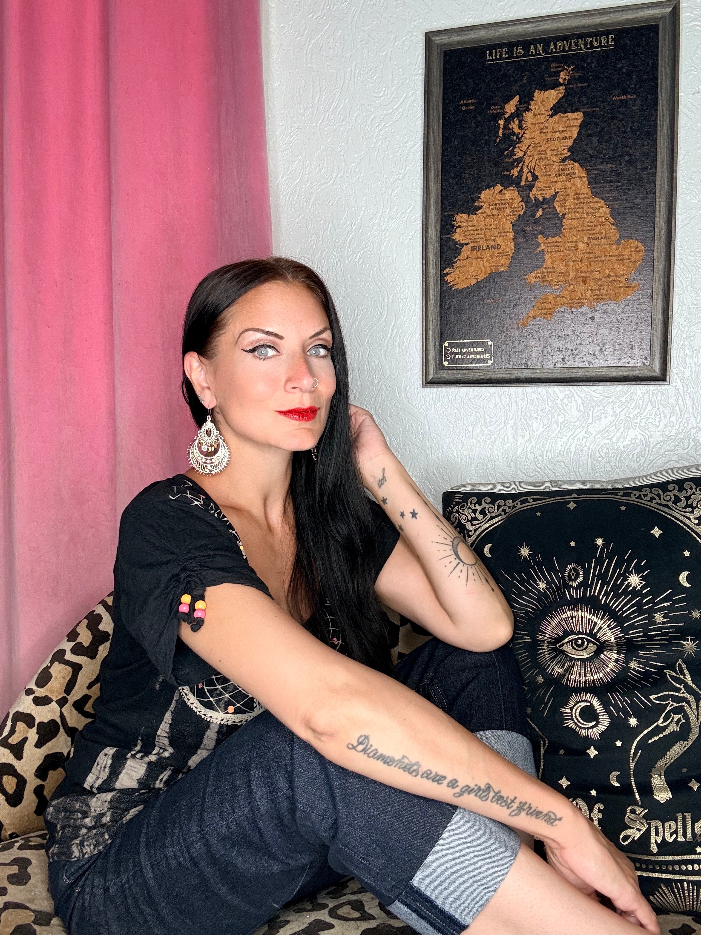 Woman with long dark hair, red lipstick, earrings, sitting on a black and gold decorative pillow, in front of pink curtain and UK map poster