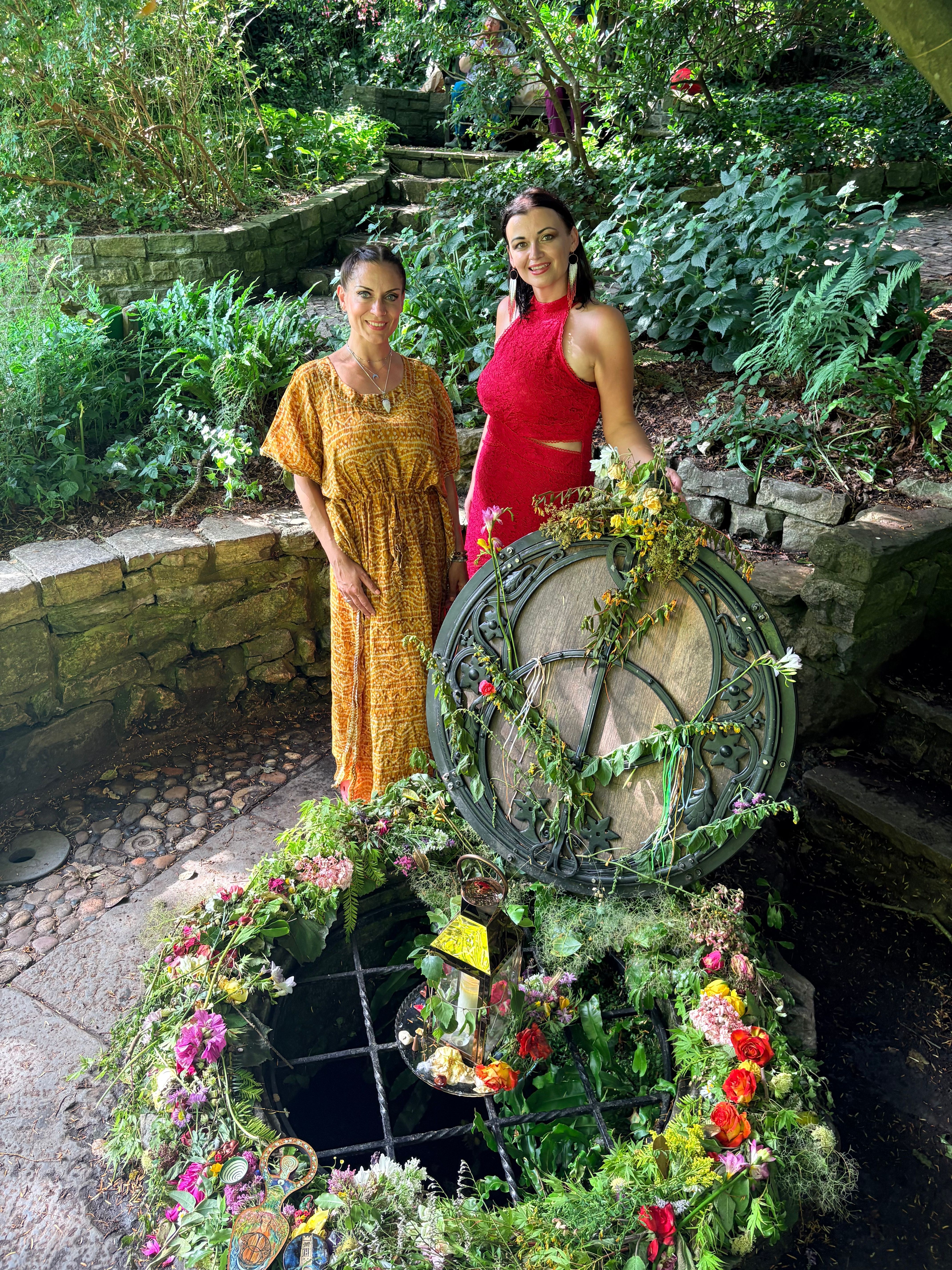 Two women stand outdoors near a bicycle decorated with flowers, surrounded by greenery and plants.