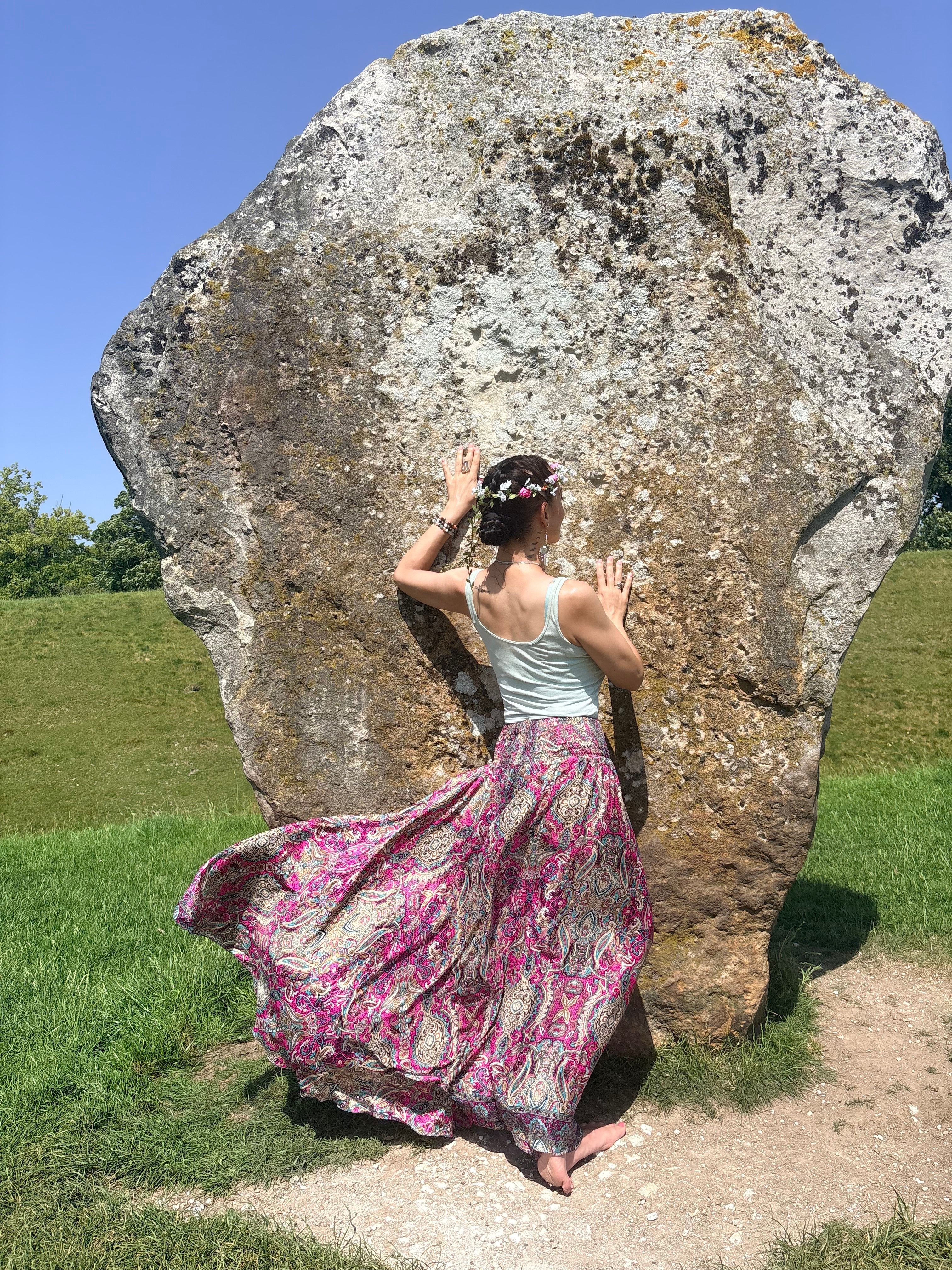 Person in a long pink and white skirt and white top leaning against a large rock outdoors.