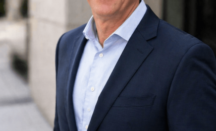 Man in a dark suit and light blue shirt standing outdoors, smiling, with a building in the background.